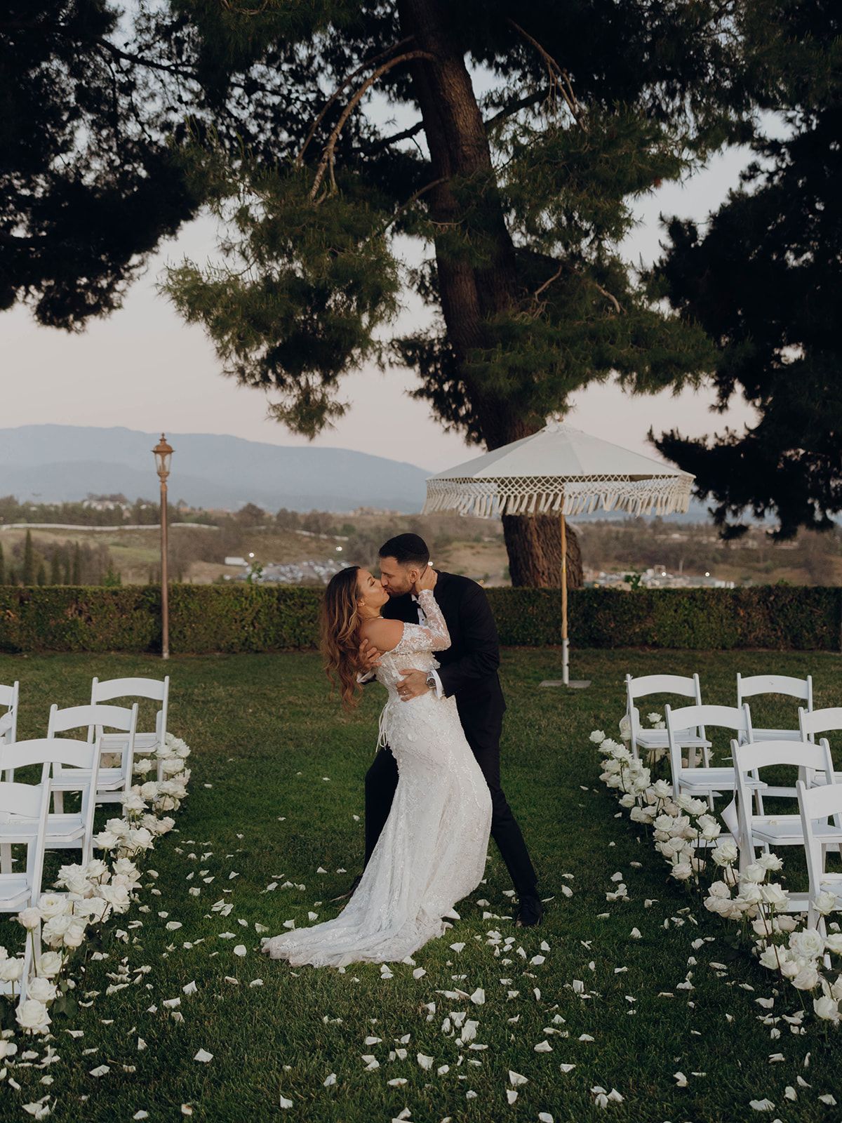 A romantic wedding ceremony setup outdoors with white chairs and flower petals lining the aisle beneath tall trees.