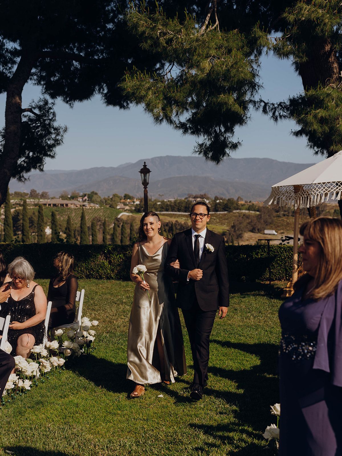 Wedding couple walks down grassy aisle at outdoor mountain venue with decorative lamp post and guests in view during sunset.