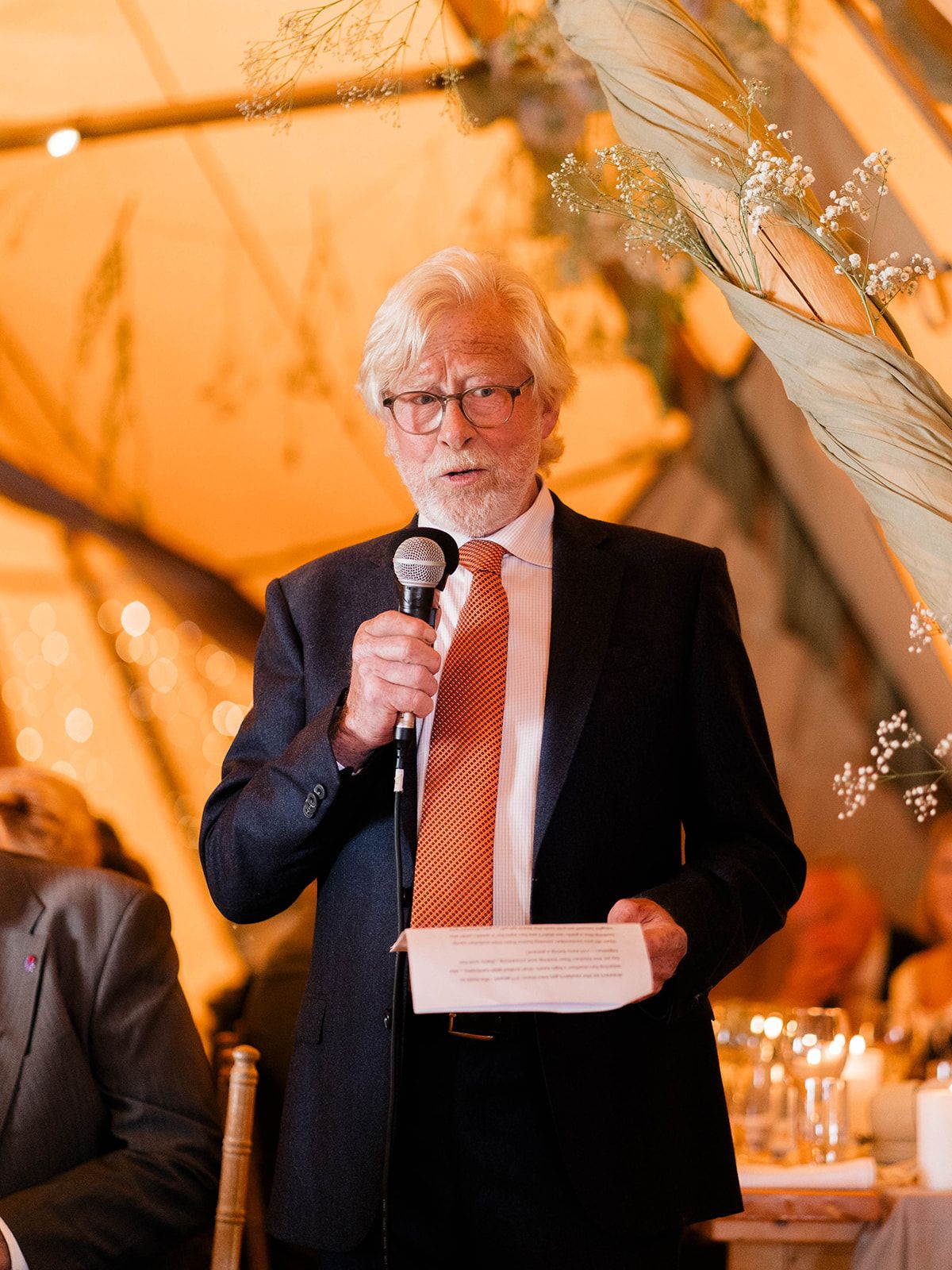 A speaker in a navy suit and orange tie delivers remarks at a formal event under warm tent lighting.