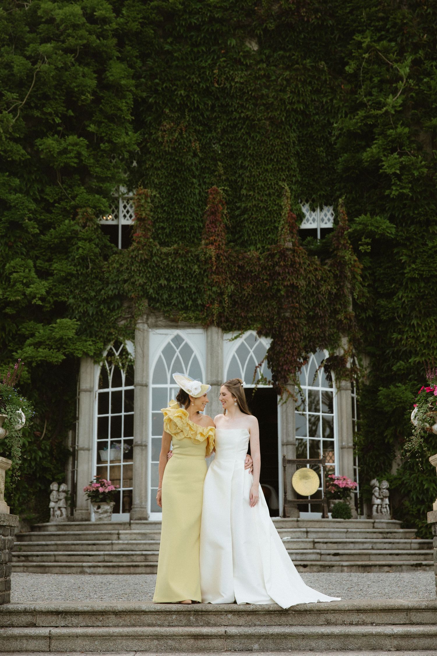 Two people in formal wear stand on stone steps in front of an ivy-covered Gothic-style building.