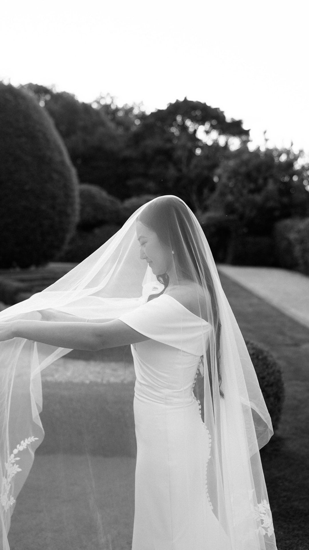 Artistic black and white photo of a bride adjusting her veil in a garden setting.