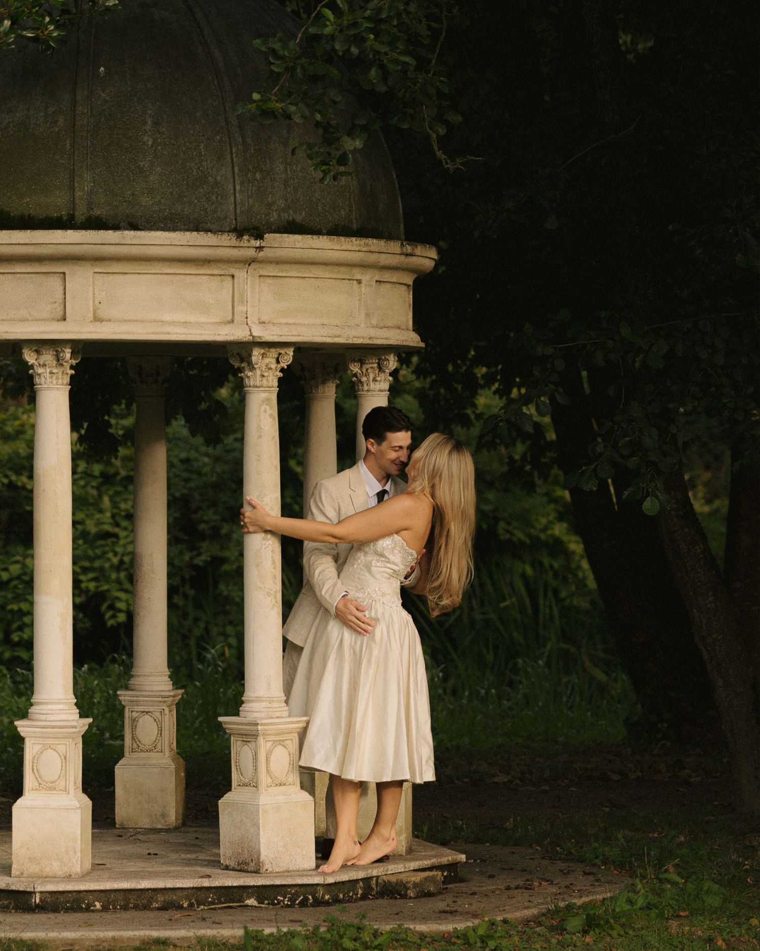 Couple embracing and kissing under a classical white gazebo surrounded by lush green trees.