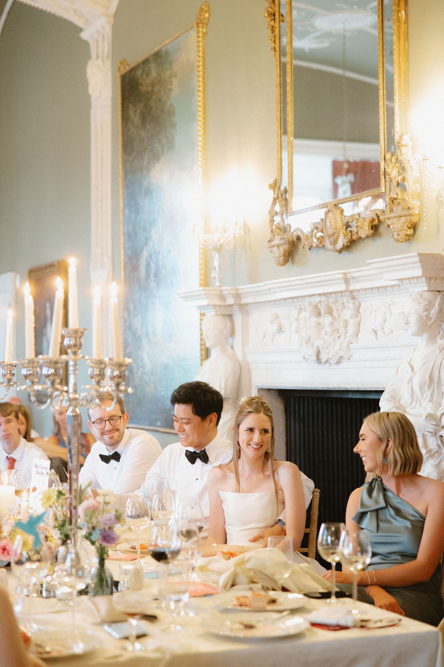 Elegant dinner party guests seated at a long table with ornate gold mirrors and candles in a luxurious dining room.