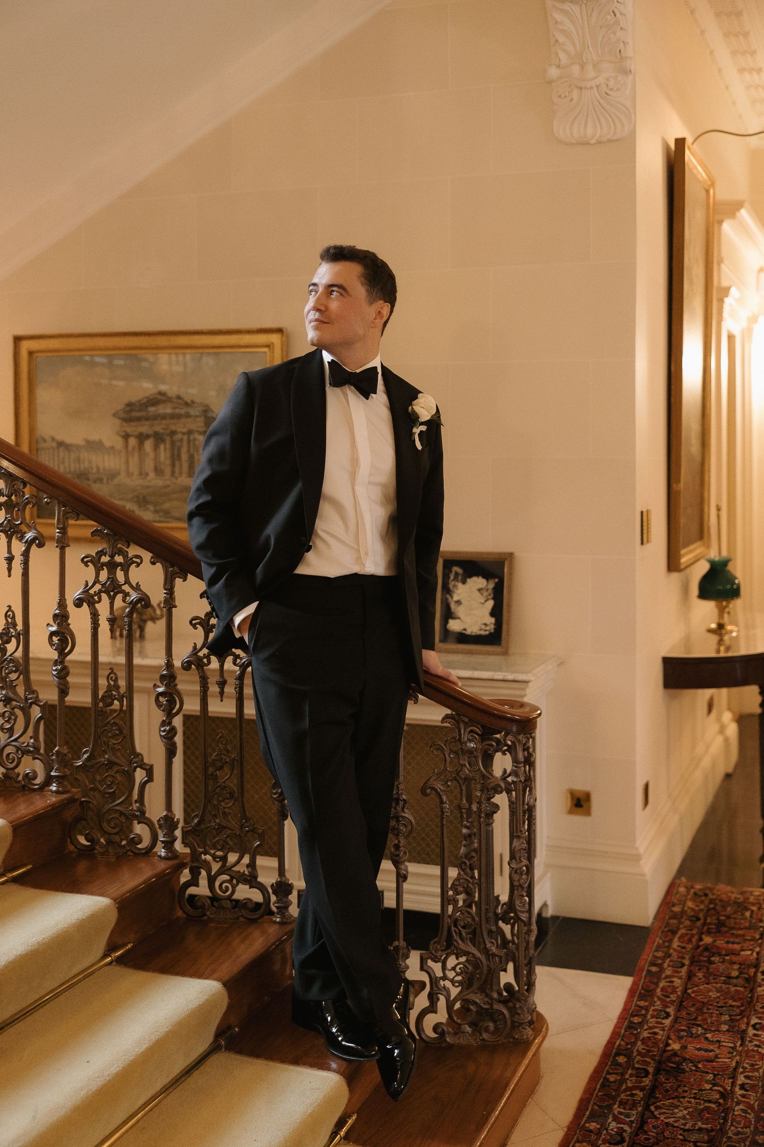 A groom in a black tuxedo poses on an elegant staircase with ornate iron railings in a vintage-style setting.
