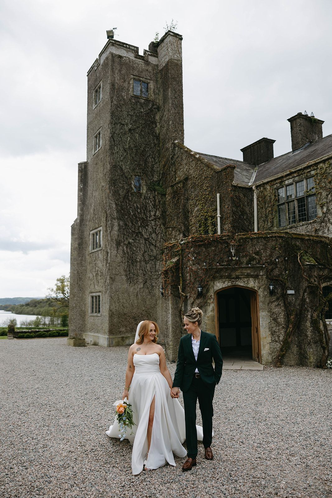 A couple walks together outside a historic stone castle with gray cloudy skies in Ireland.