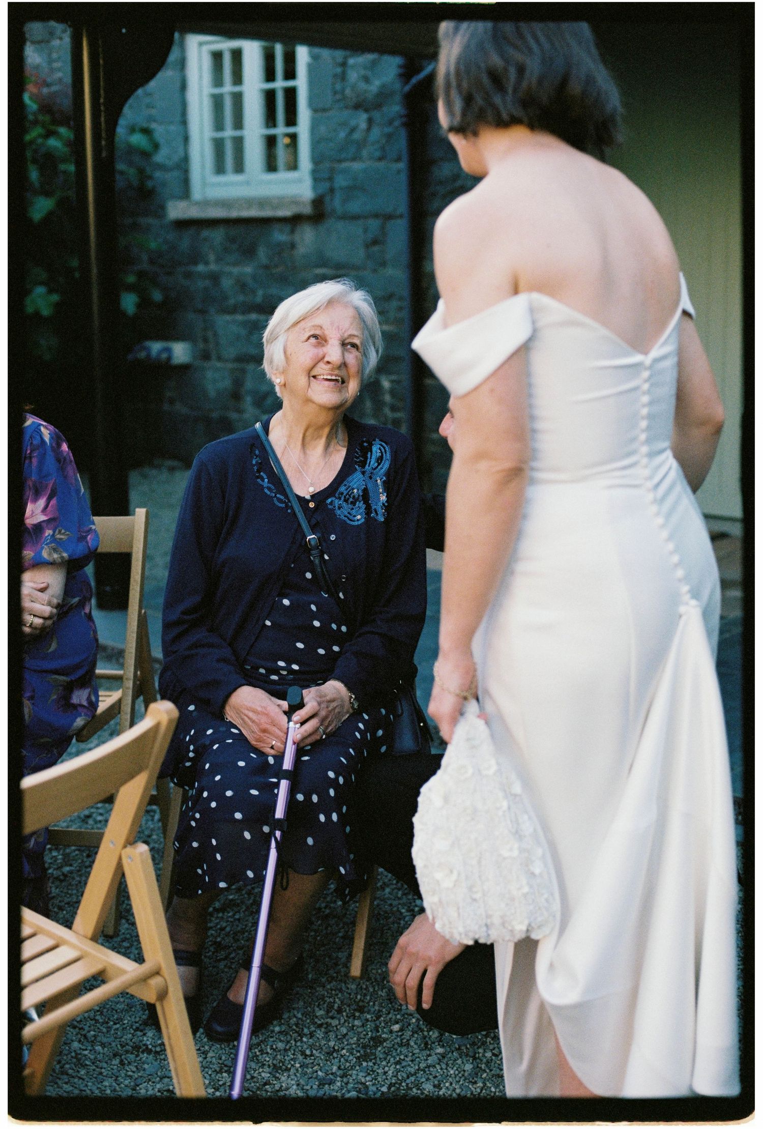 Elderly woman with cane smiling up at bride in white off-shoulder wedding gown outdoors.