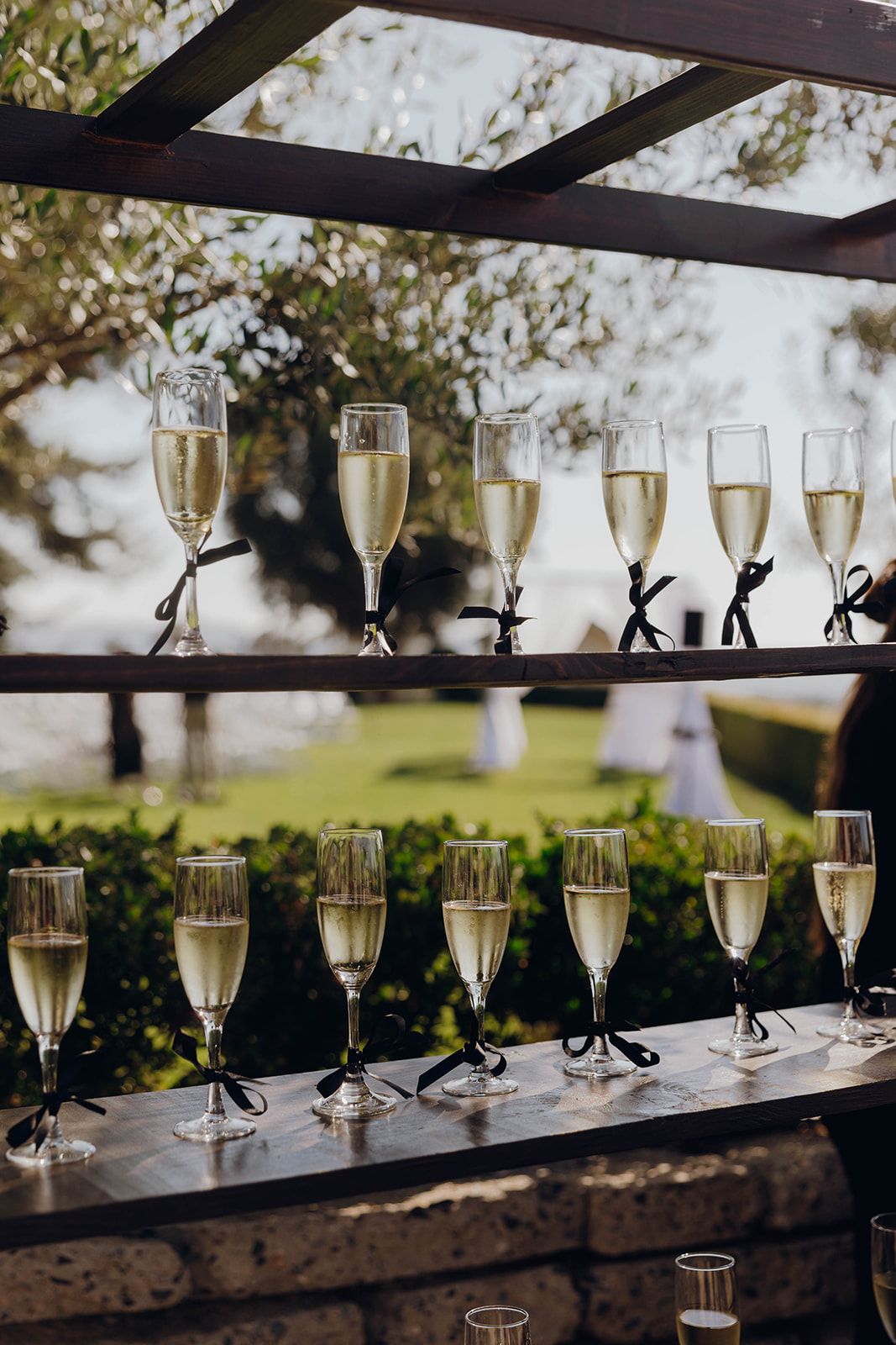 Rows of elegant champagne flutes decorated with black ribbons displayed on rustic wooden shelves at an outdoor event.