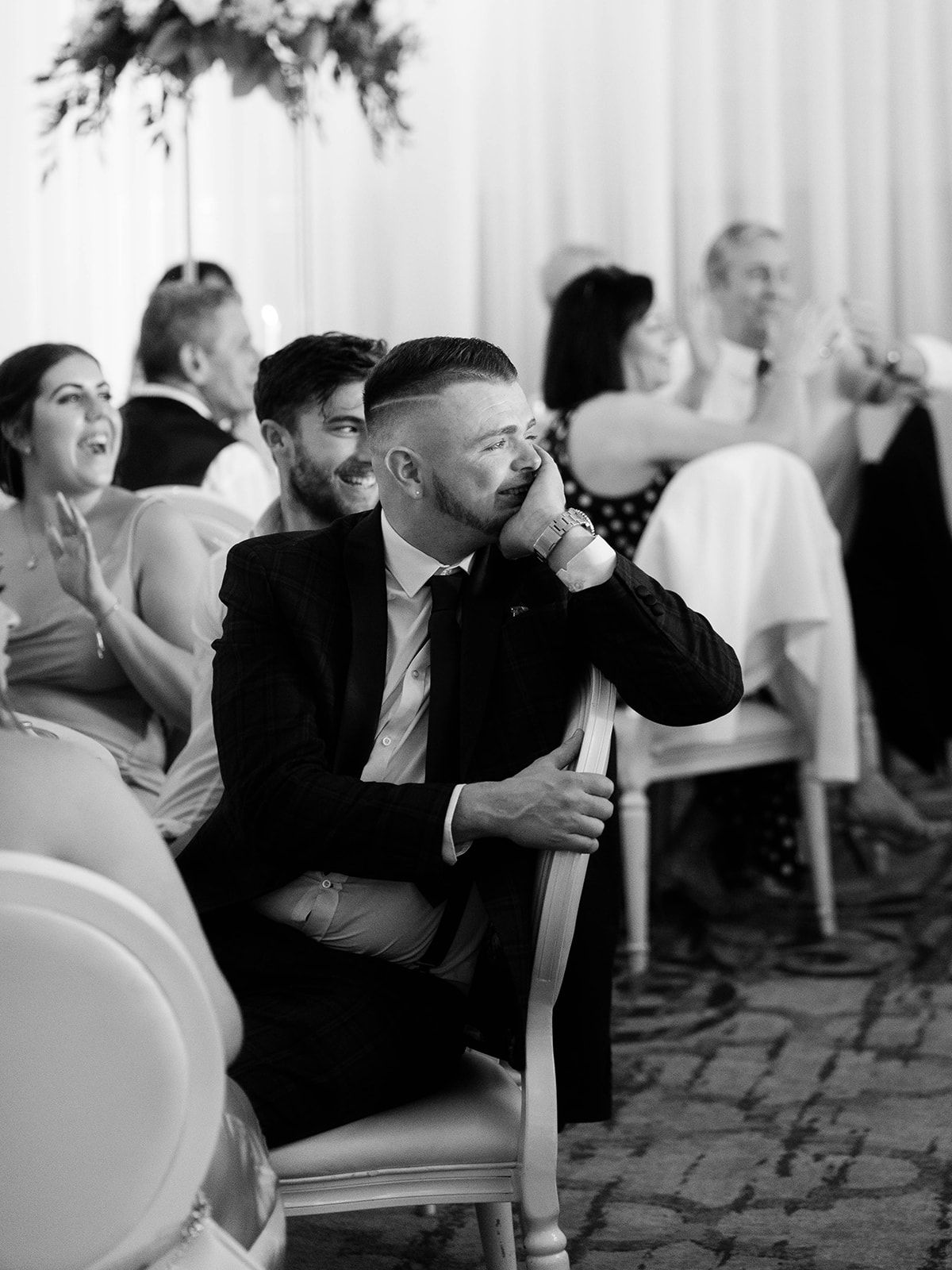 Wedding guests laugh and react emotionally during what appears to be a reception speech or toast at an indoor venue.