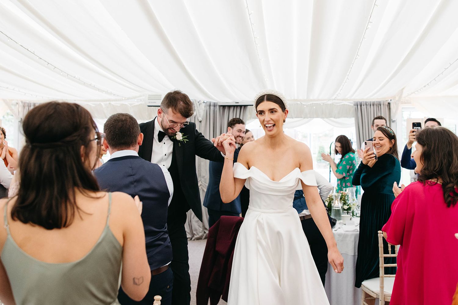 Joyful celebration at an elegant wedding reception inside a white tent with guests mingling and dancing.
