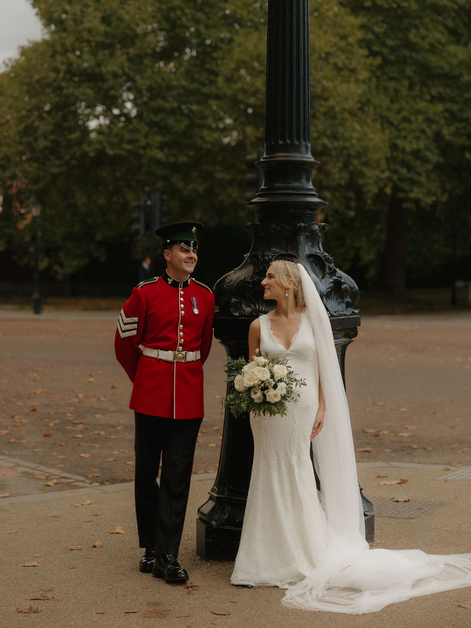 A military guard in red uniform walks alongside a bride in a flowing white wedding gown in a park setting.