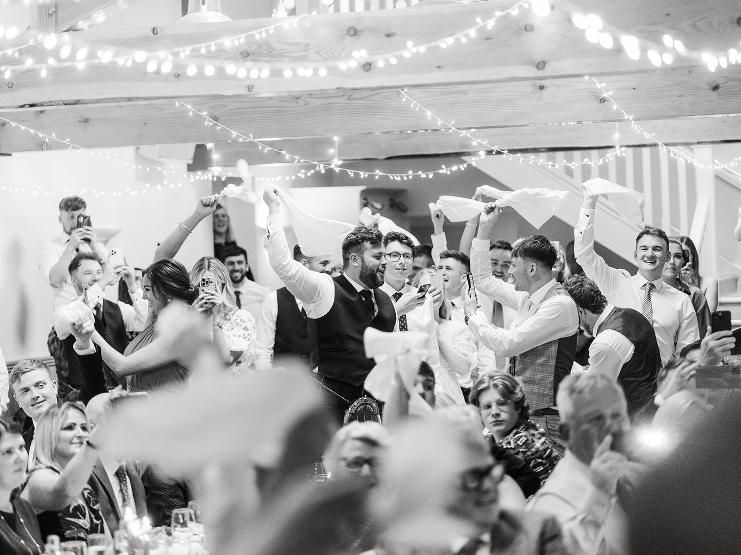 A celebratory wedding reception scene with guests raising their glasses during a toast under decorative hanging lights.