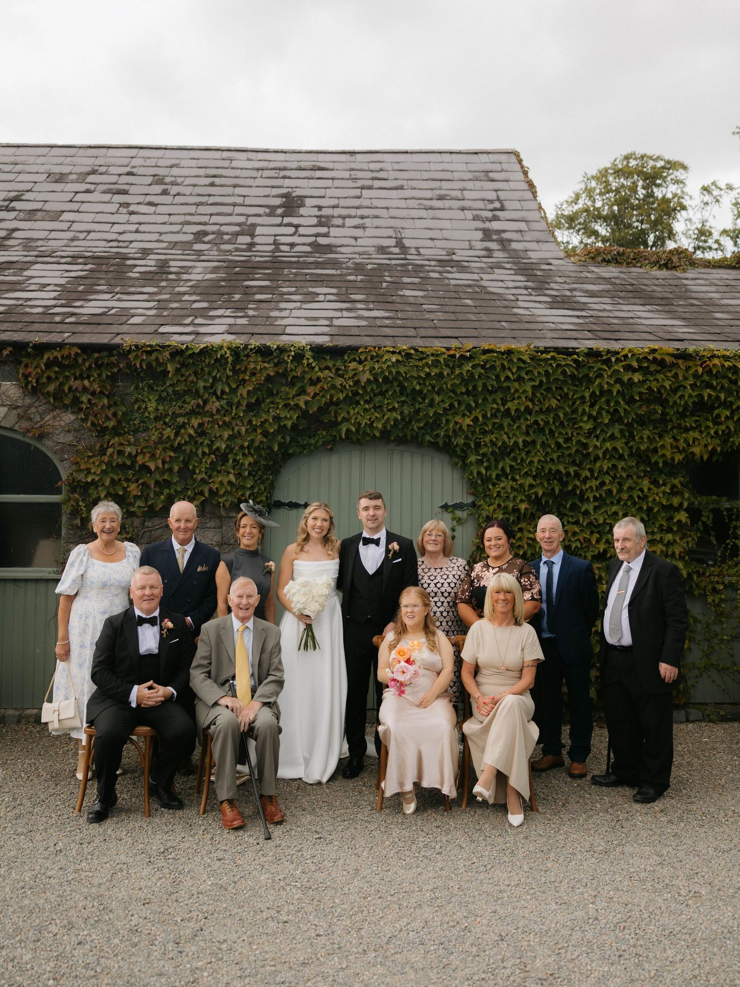 A group gathers for a formal photo outside an ivy-covered building with a gray shingled roof.