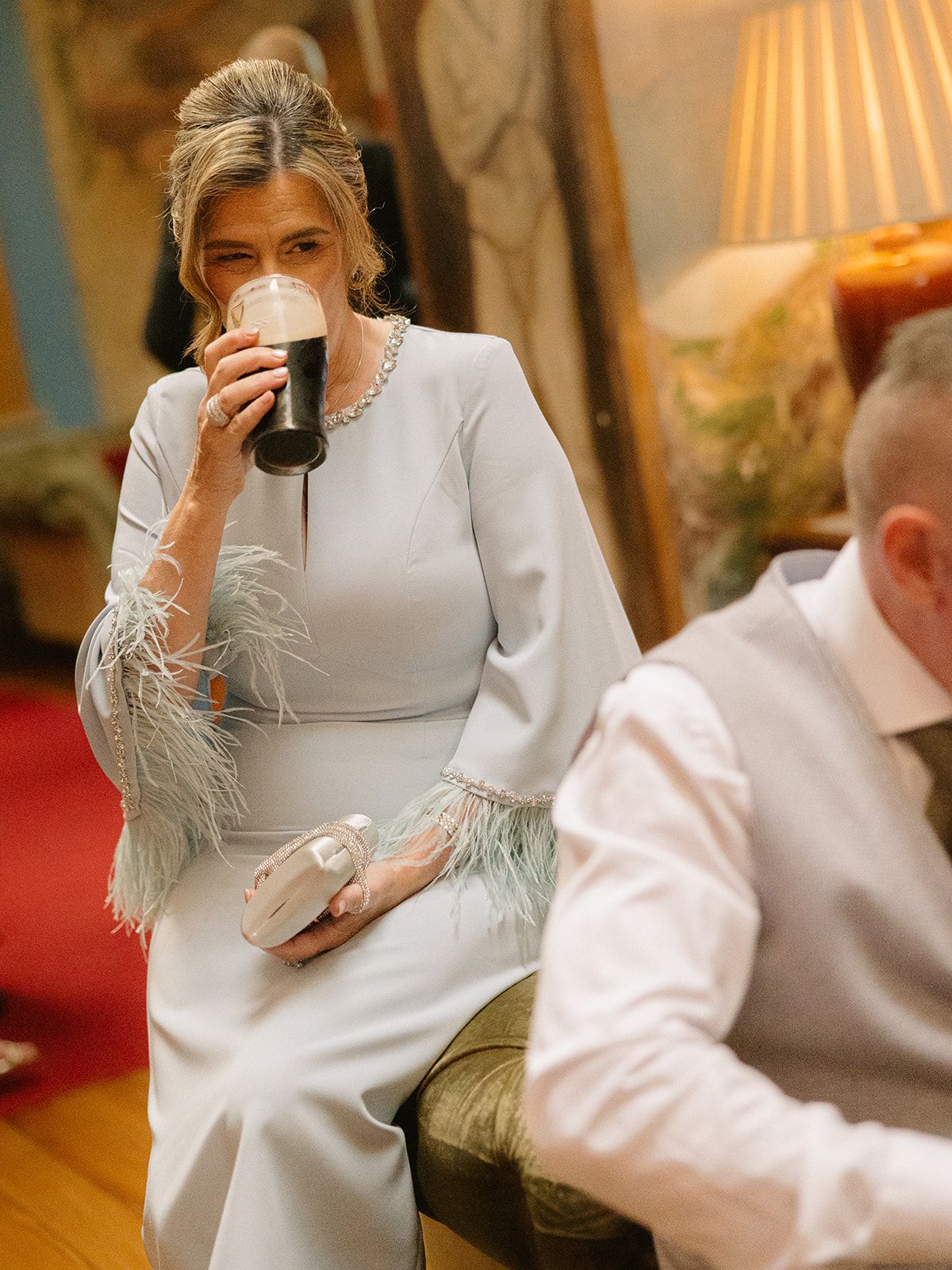 A person in a white dress sits elegantly while holding a cup in a warmly lit room with classic decor.