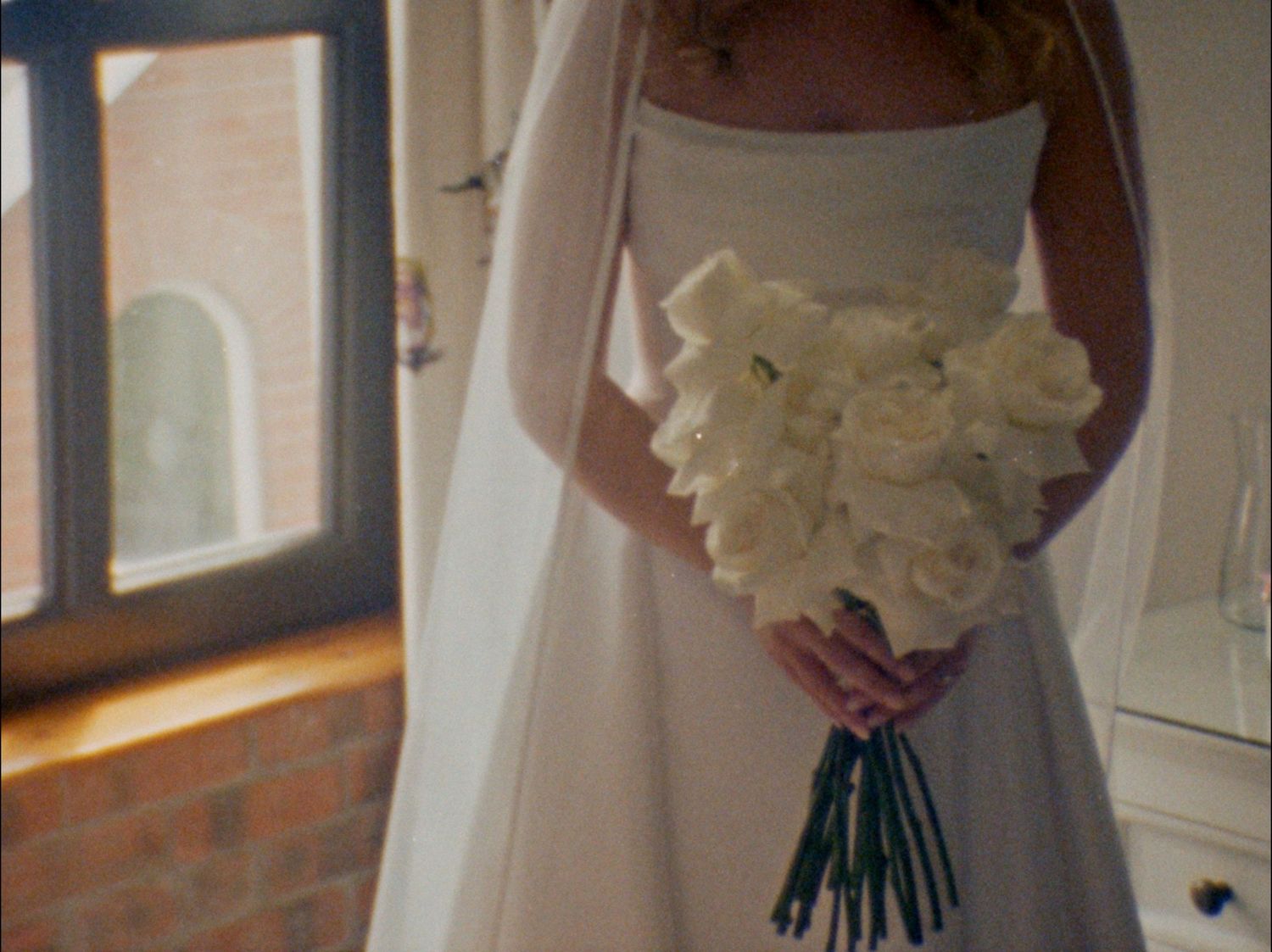 A bride in a white strapless wedding dress holds a bouquet of white flowers while standing near a window.