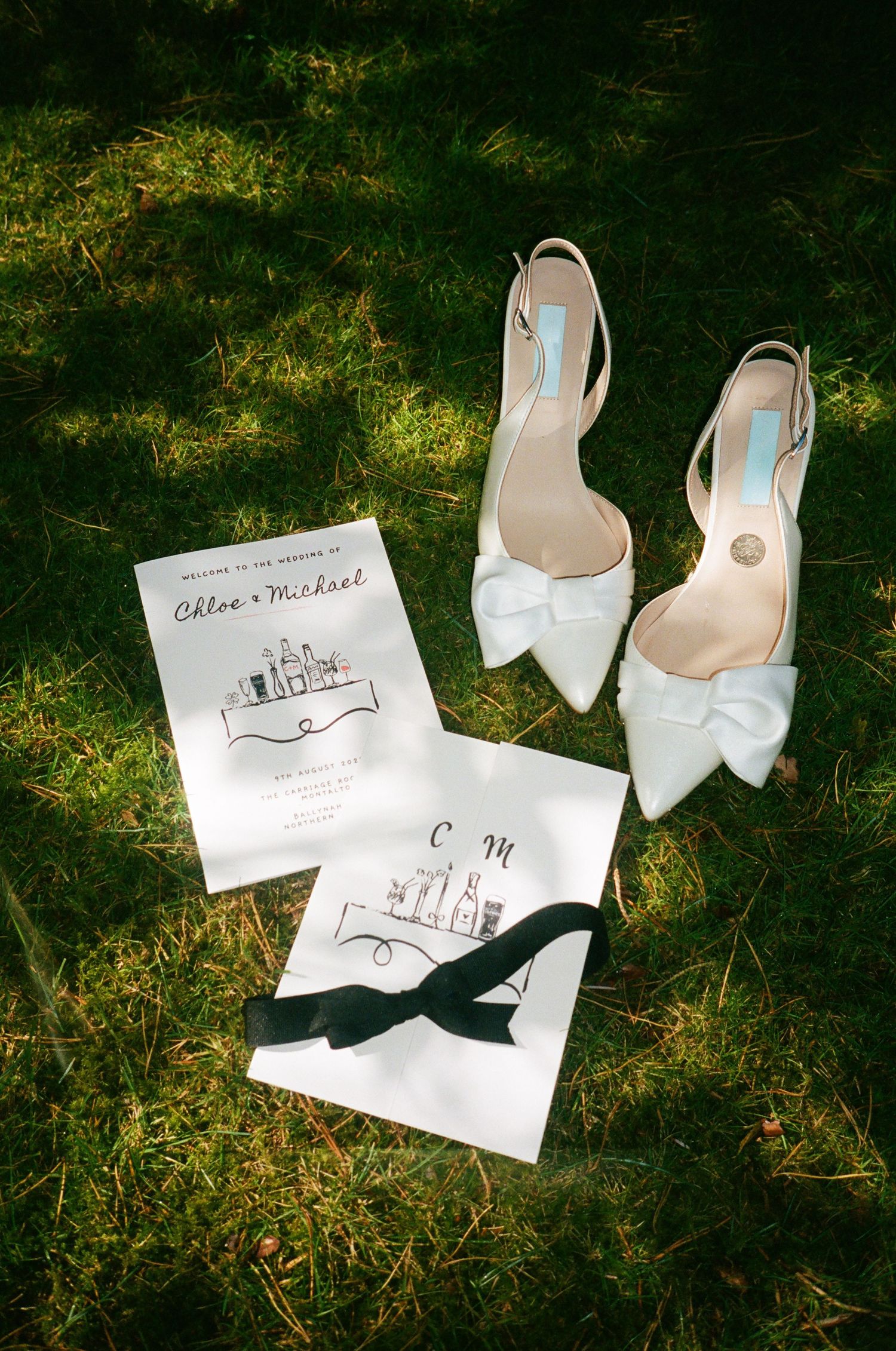 White bridal shoes and wedding invitation cards arranged on grass.