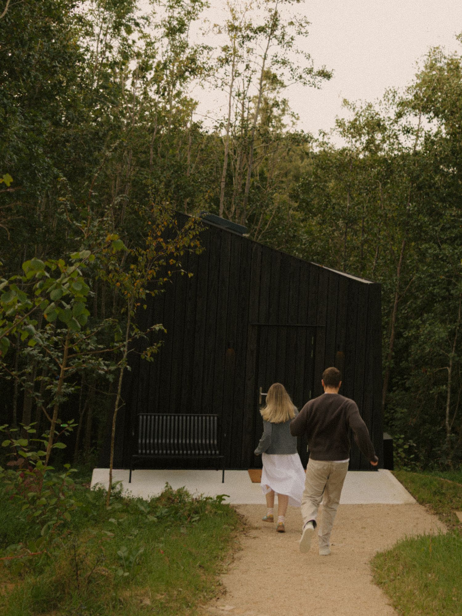 Couple walking toward a dark barn-style cabin surrounded by lush green forest trees on a gravel path.