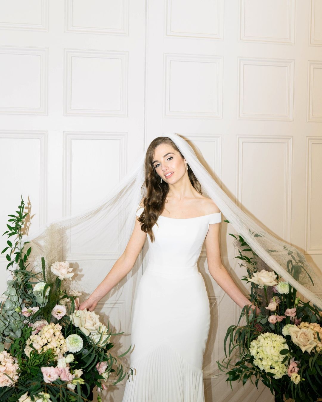 Elegant off-shoulder white wedding dress with flowing veil surrounded by pink and cream floral arrangements against white wall.