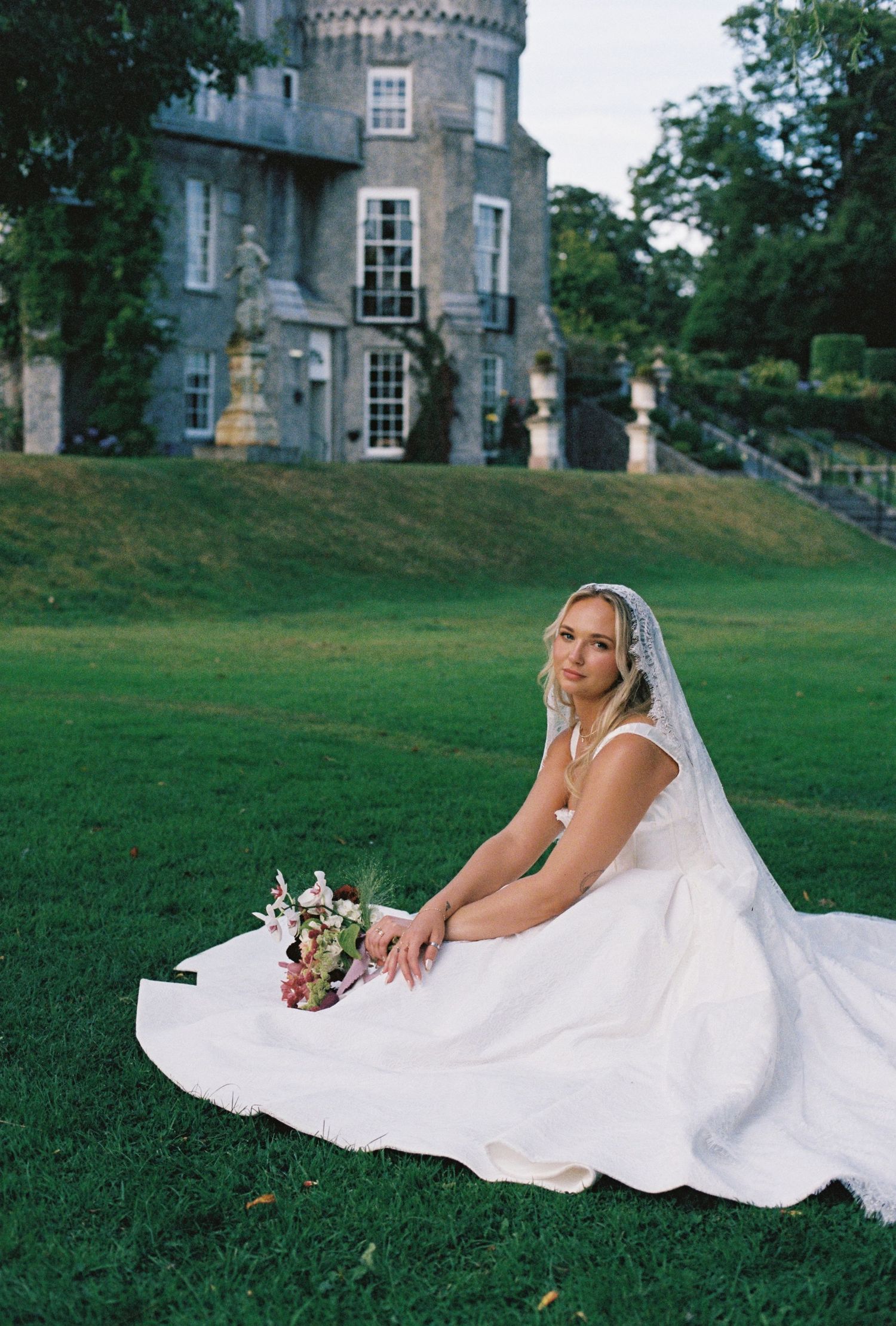 Bride in white gown and veil sits on green lawn holding bouquet before a grand stone castle.