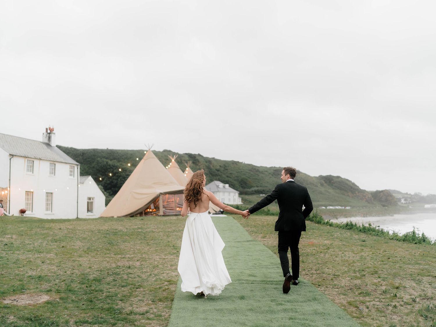 A couple runs hand in hand on a path near coastal cottages and beige tents.