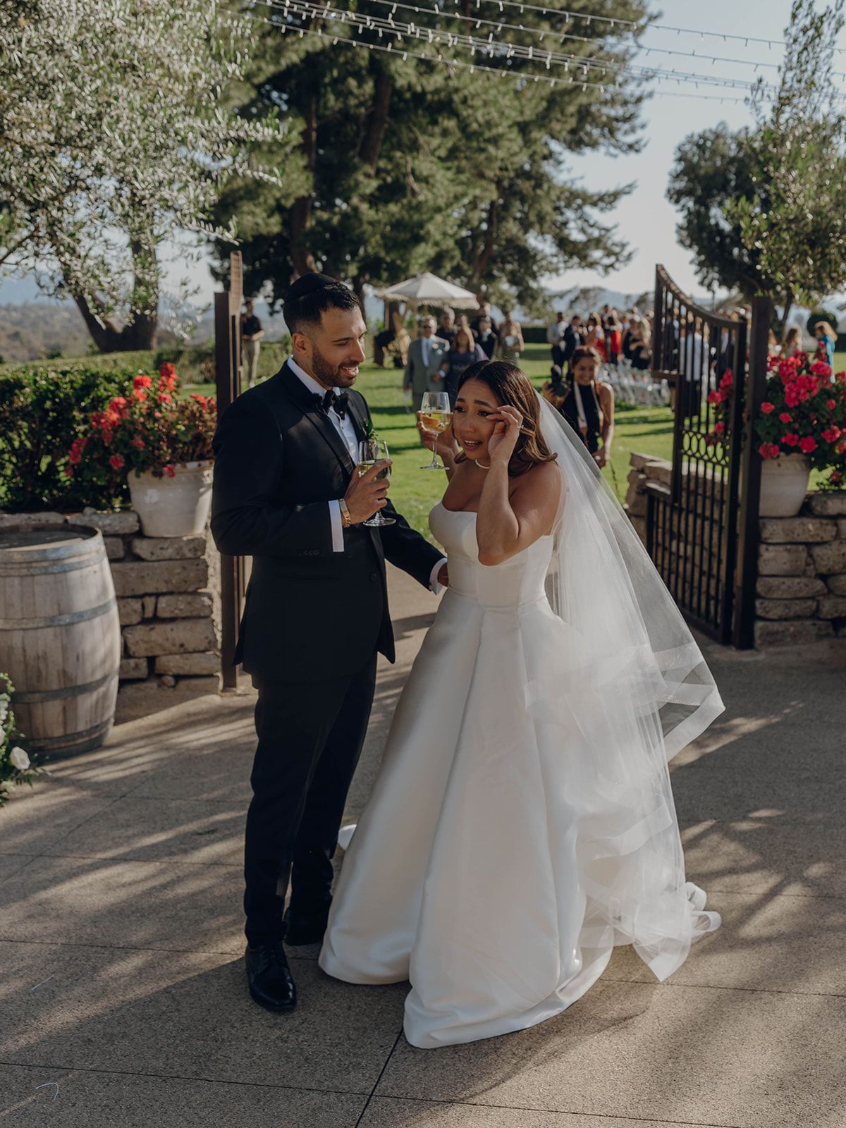 A joyful candid wedding moment between newlyweds at a sunny winery venue with decorative wine barrels and red flowers.