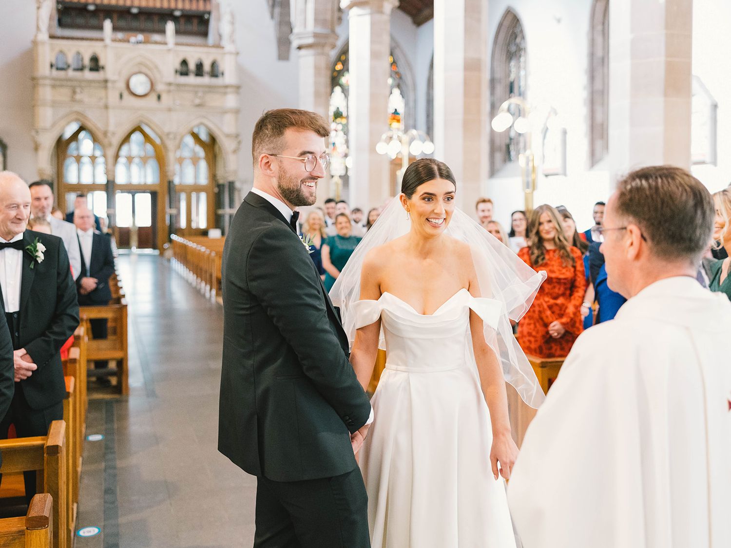 A couple exchanges vows during their wedding ceremony inside a grand cathedral with Gothic arched windows and wooden pews.