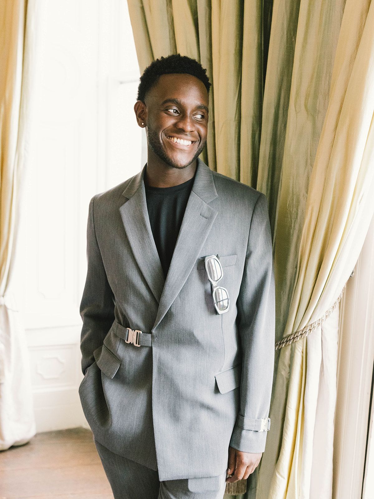 A person wearing a sleek grey suit with a black shirt smiles brightly in front of cream colored curtains.