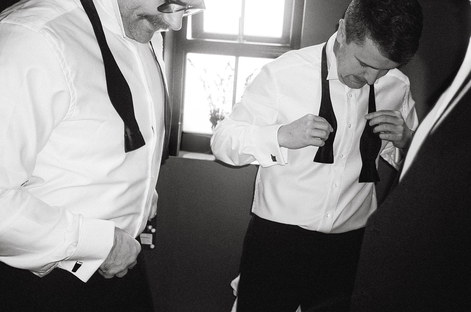 Two men in formal attire with white dress shirts and suspenders prepare their outfits in a black and white photograph.