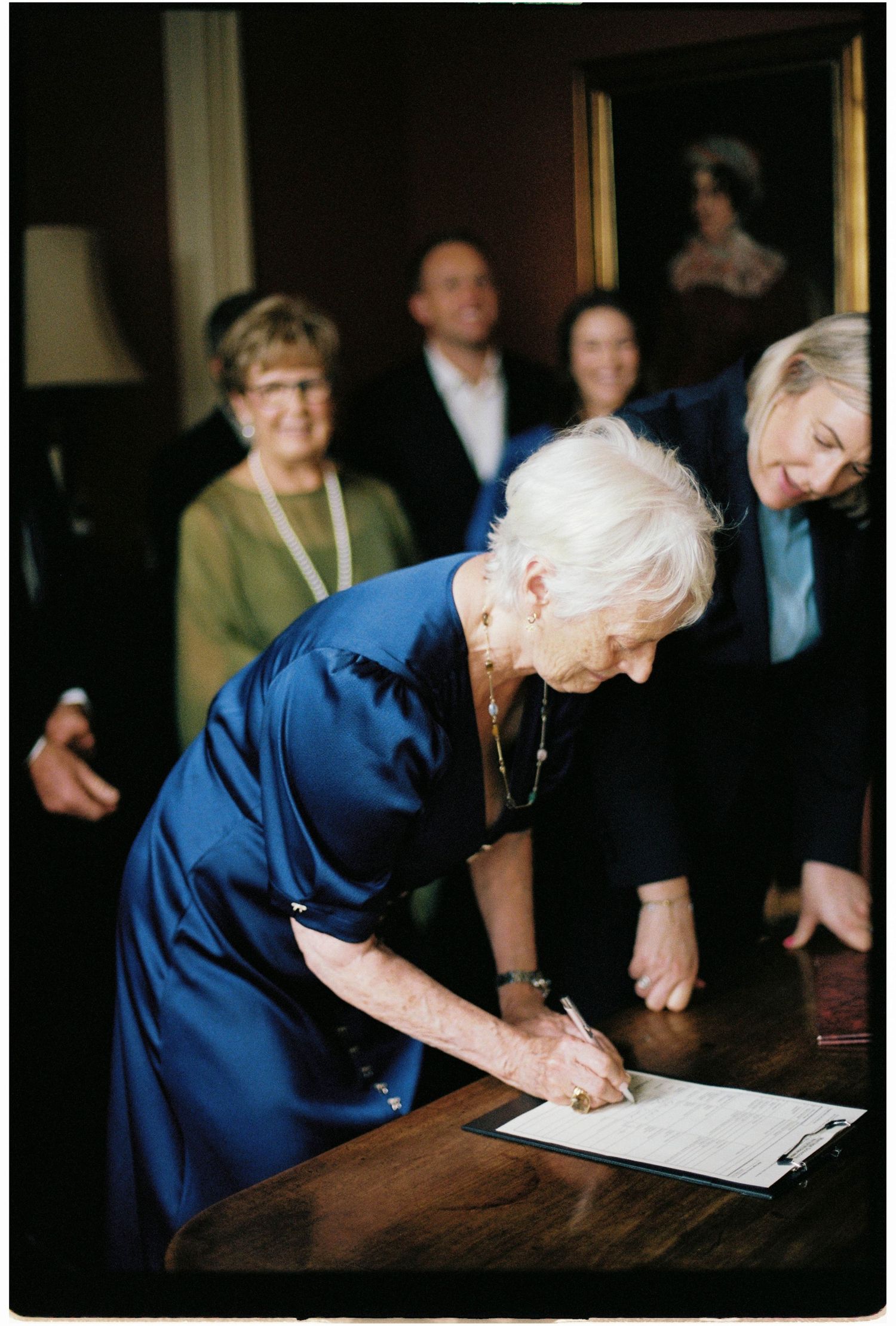 Elderly woman in blue dress signing documents at a wooden table during a wedding ceremony.