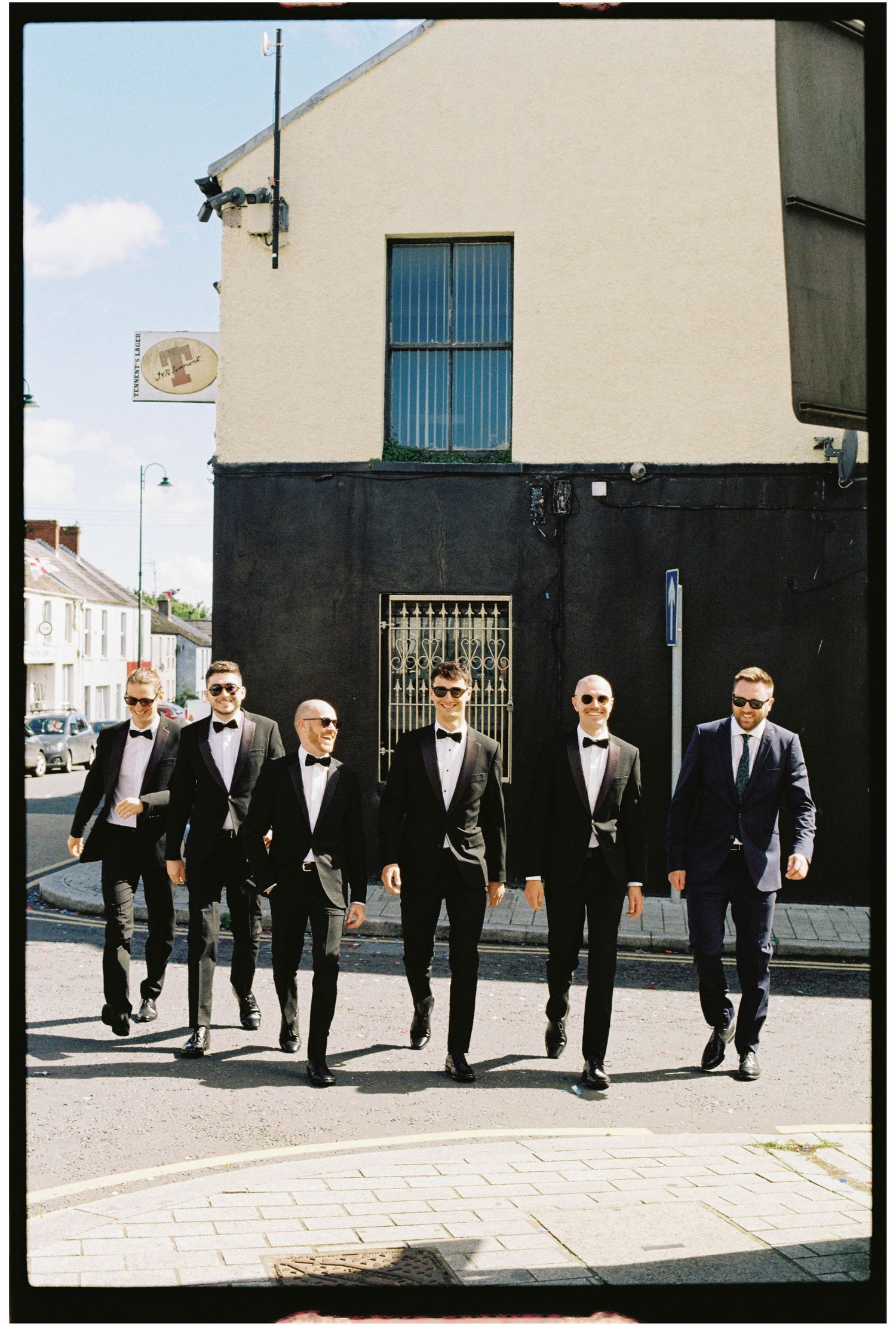 Six groomsmen in black tuxedos and bow ties walk confidently down a sunny street on a wedding day.