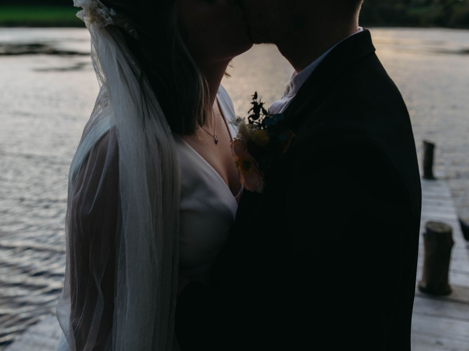 A romantic wedding couple embraces near a waterfront at sunset, with the bride's veil flowing in the breeze.
