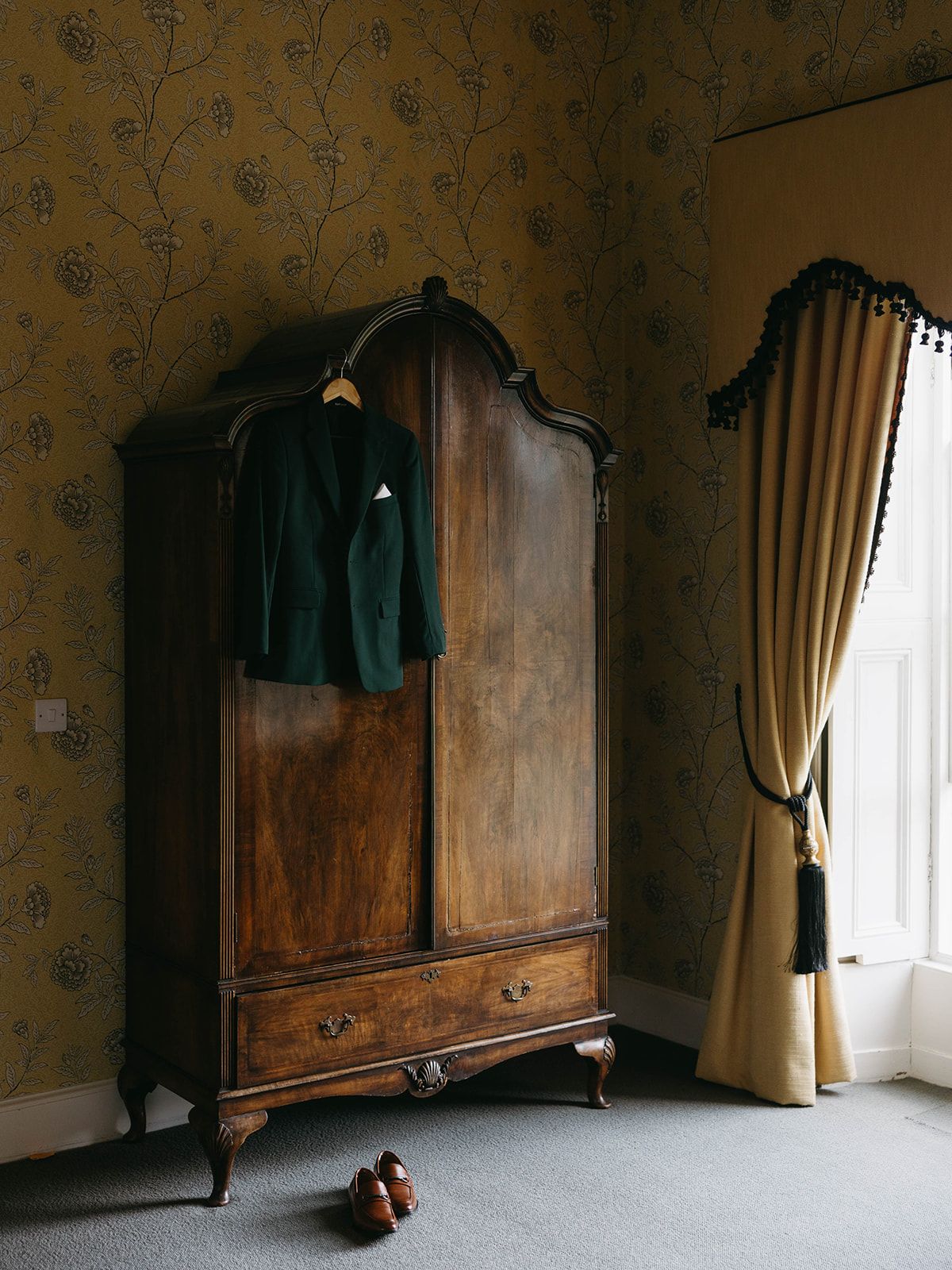 A vintage wooden wardrobe stands against floral wallpaper in a dimly lit room with beige curtains and brown shoes on the floor.
