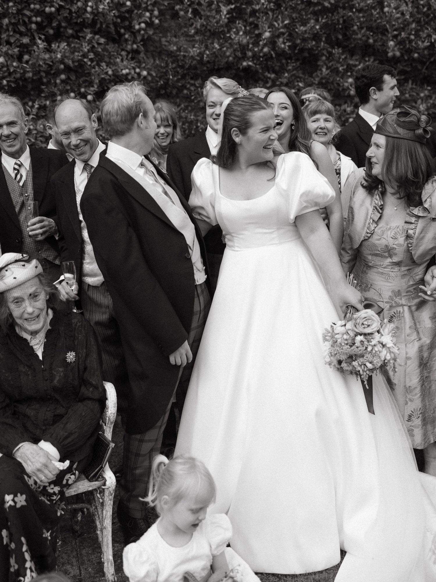 A black and white wedding photo showing a bride in a classic white dress surrounded by wedding guests outdoors.