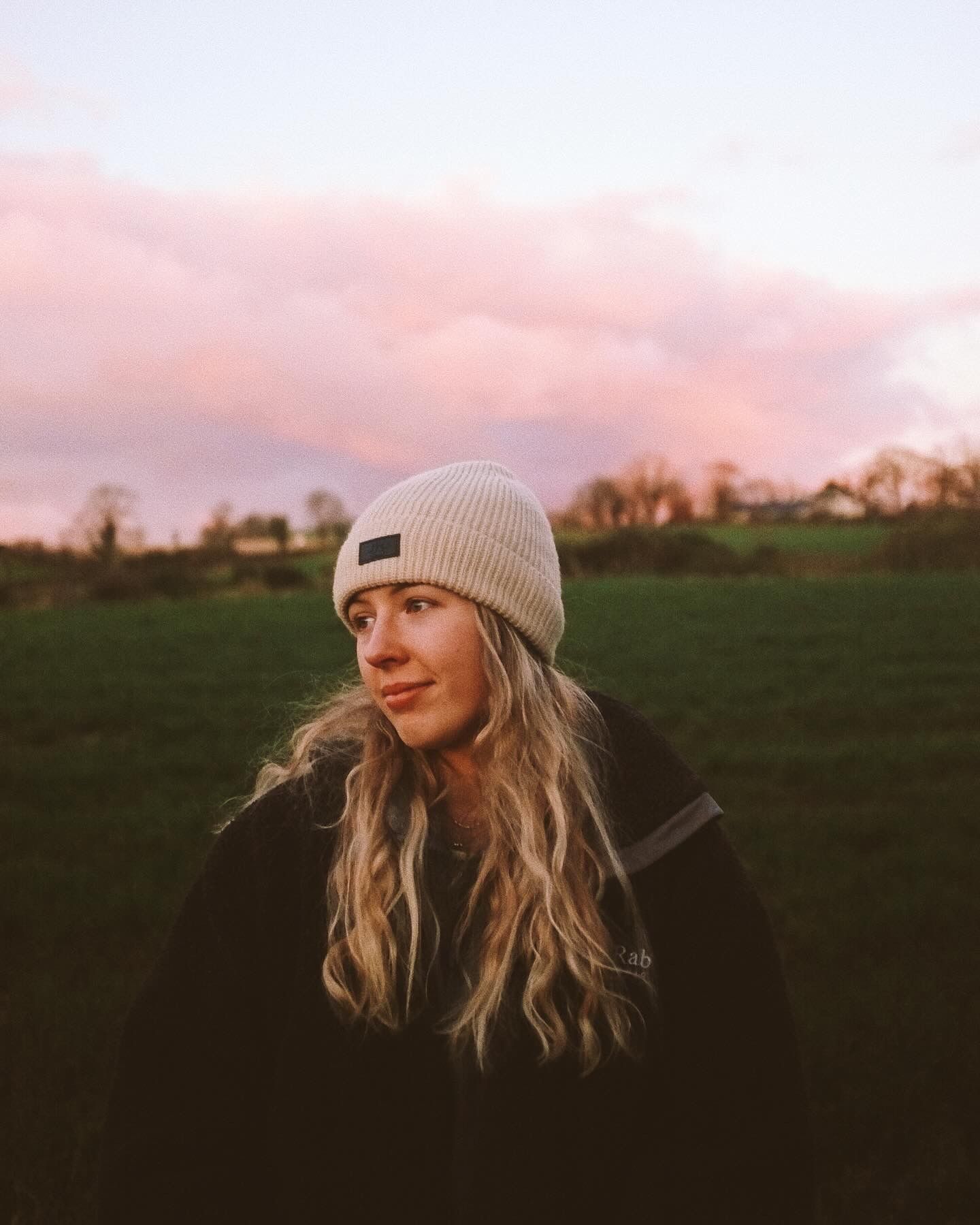 Person wearing a beige beanie and dark jacket stands in a field during sunset with pink clouds in the sky.
