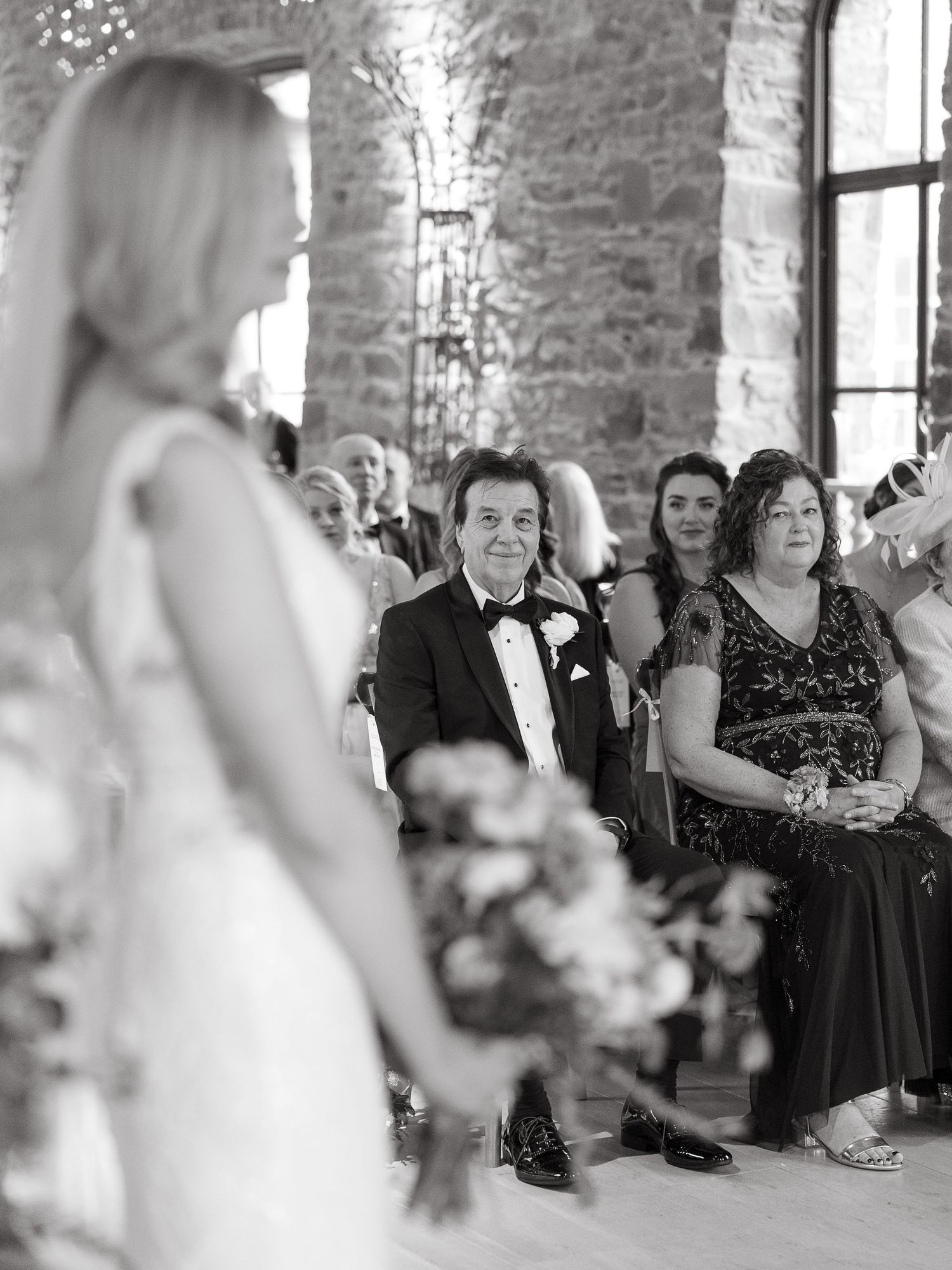 Black and white wedding photo of guests watching bride walk down the aisle in a stone venue.