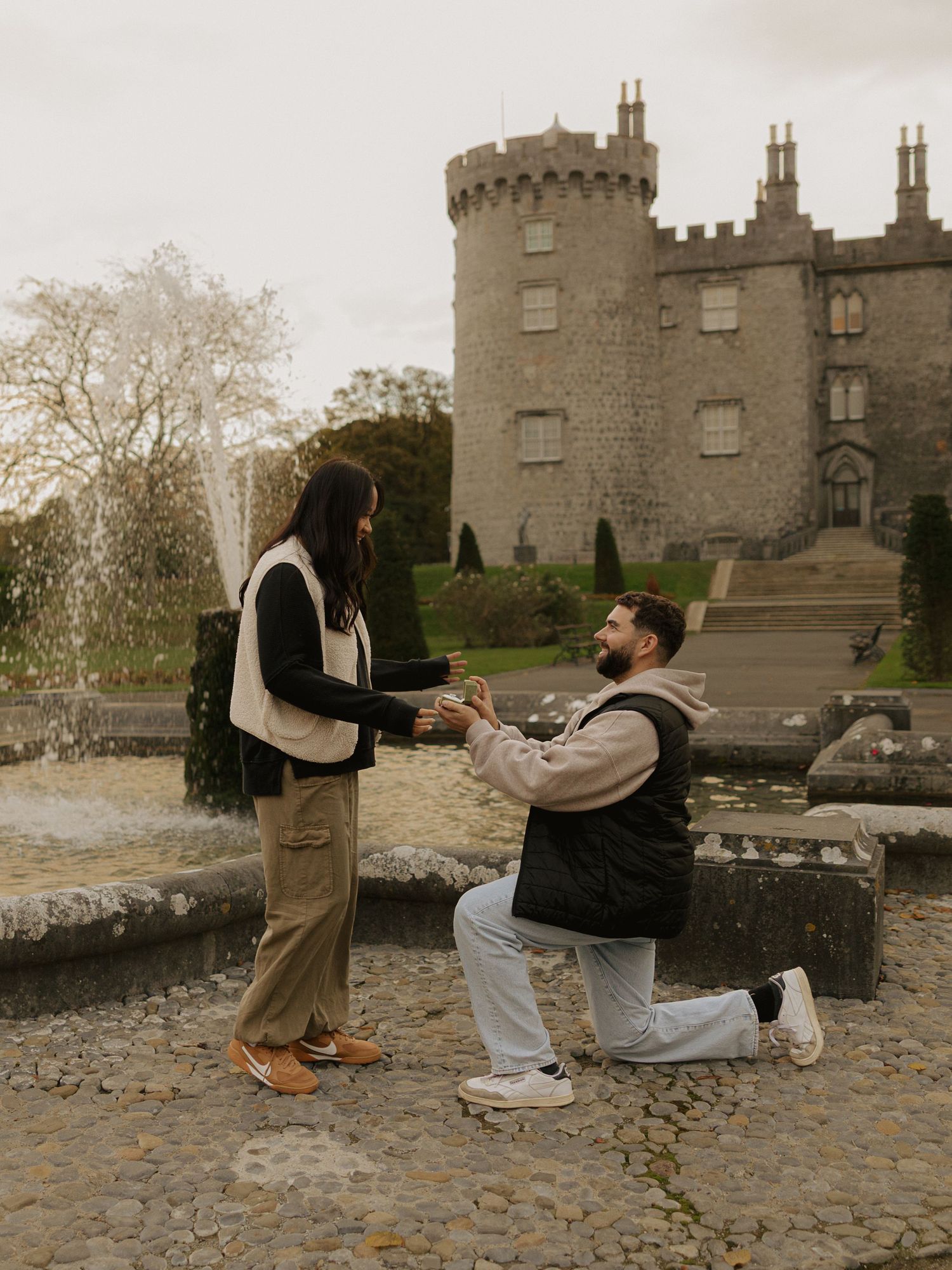 A man proposes on one knee to a woman in front of a historic stone castle with a fountain in Ireland.