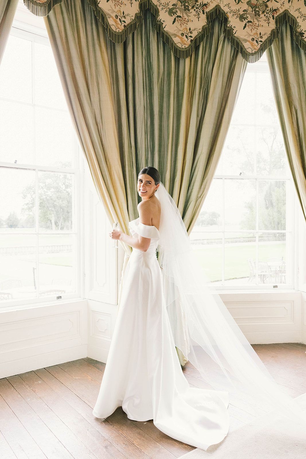 Elegant wedding dress with off-shoulder design and flowing train photographed against sage green draped curtains in bright room.