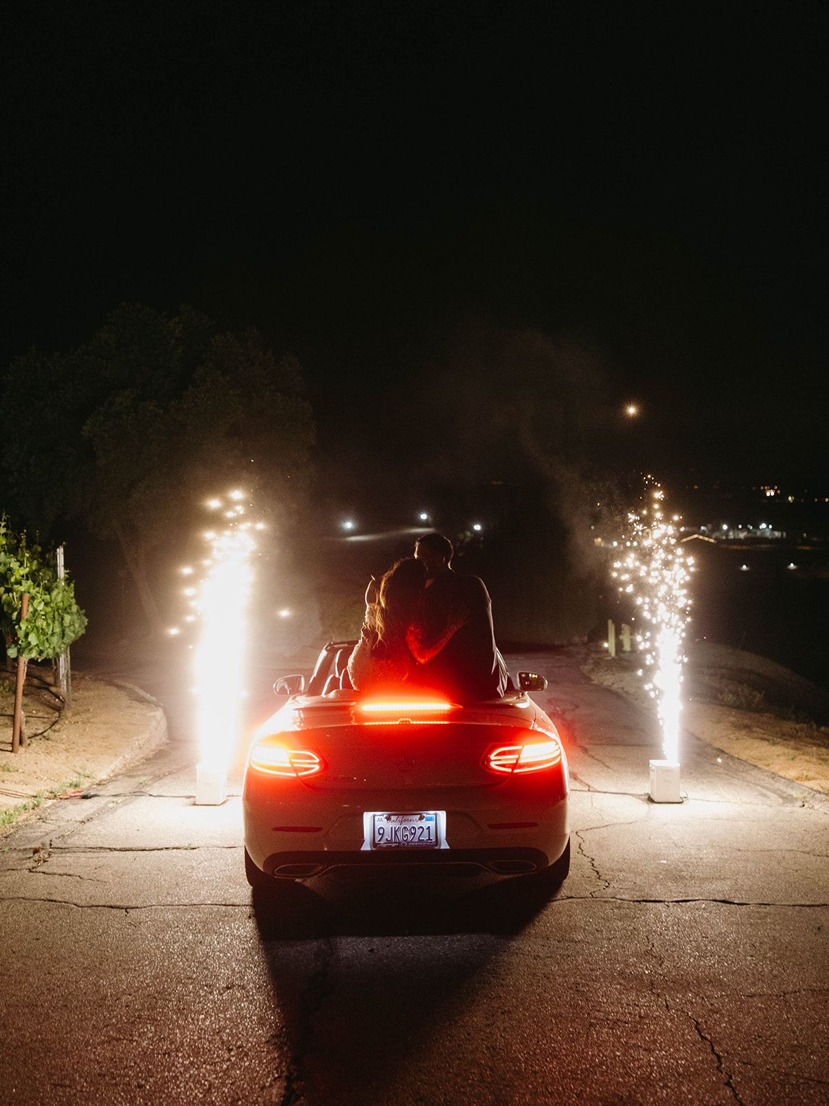 A car sits at night with brake lights glowing and sparkler fireworks creating bright light trails on both sides.
