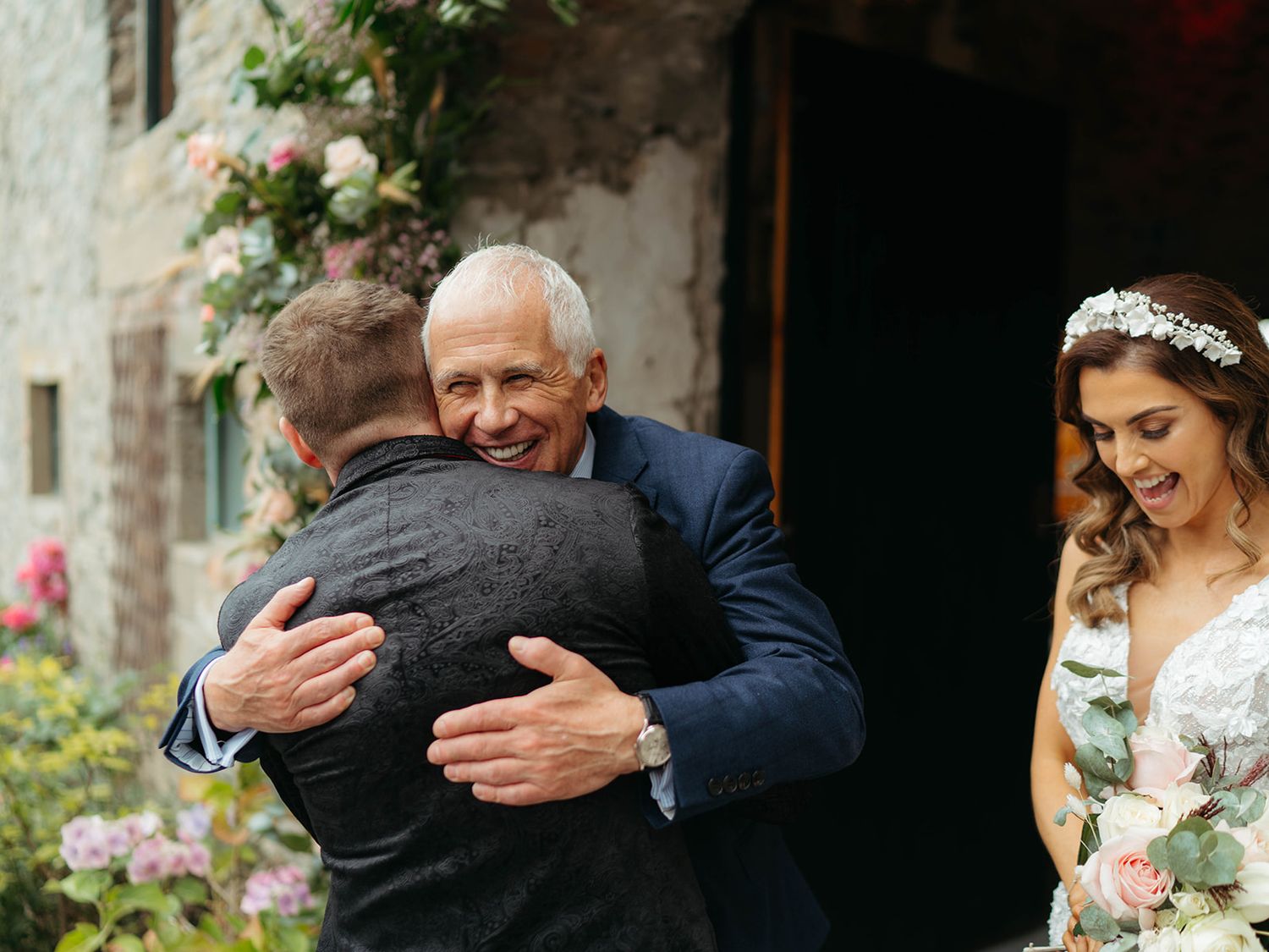 Emotional wedding scene with family members embracing at stone building entrance while bride looks on with bouquet.
