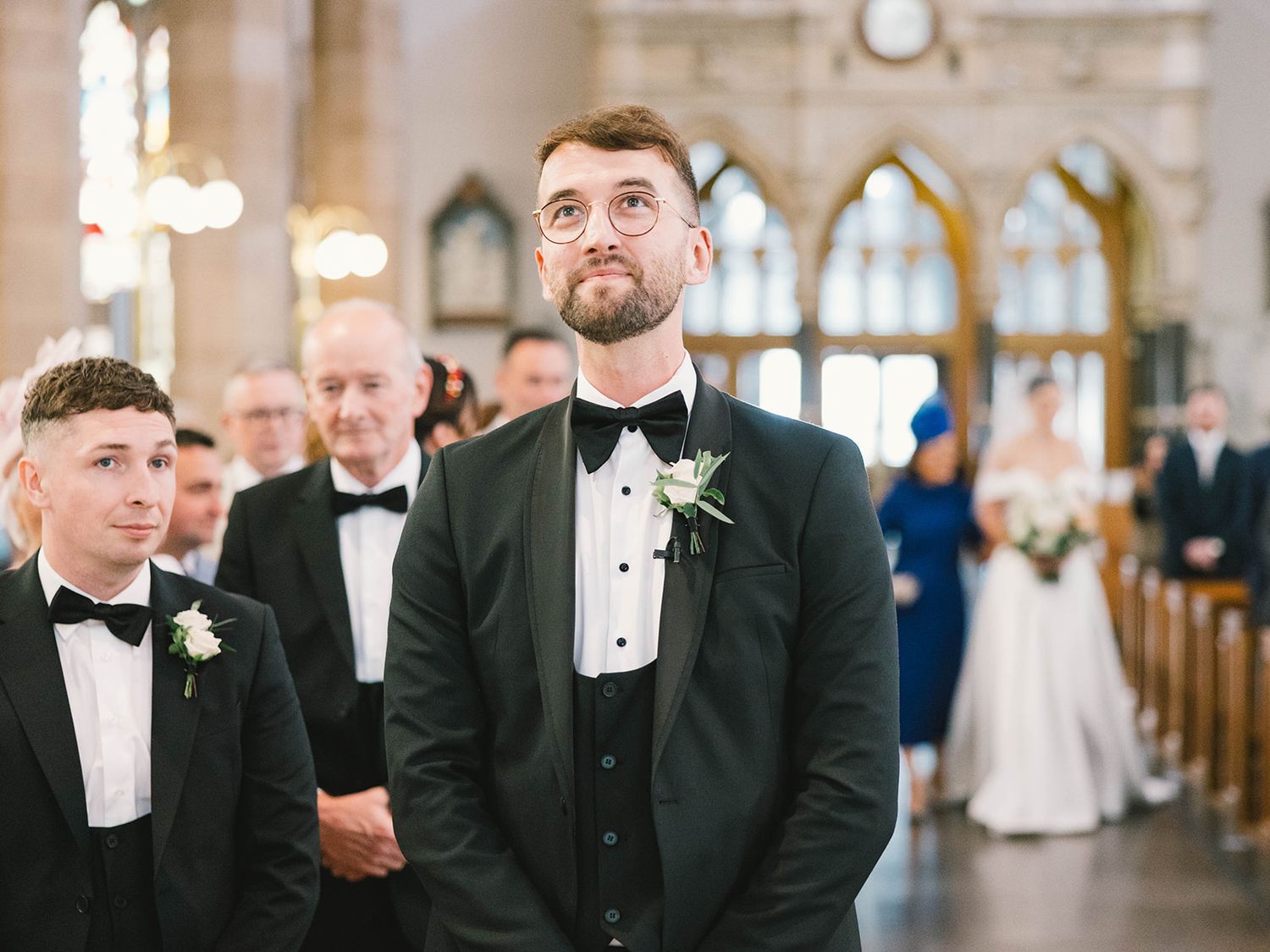 Wedding party members in formal attire stand in a beautiful church with stained glass windows and gothic architecture.