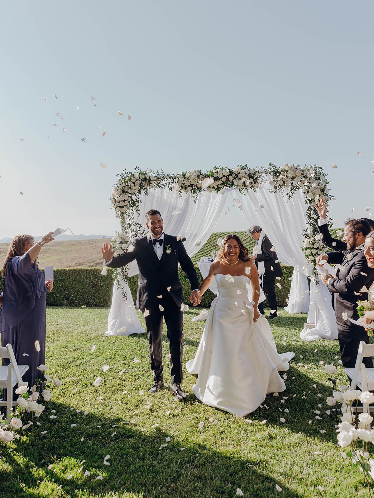 An outdoor wedding ceremony with white floral arch and guests throwing petals as couple celebrates under blue sky.