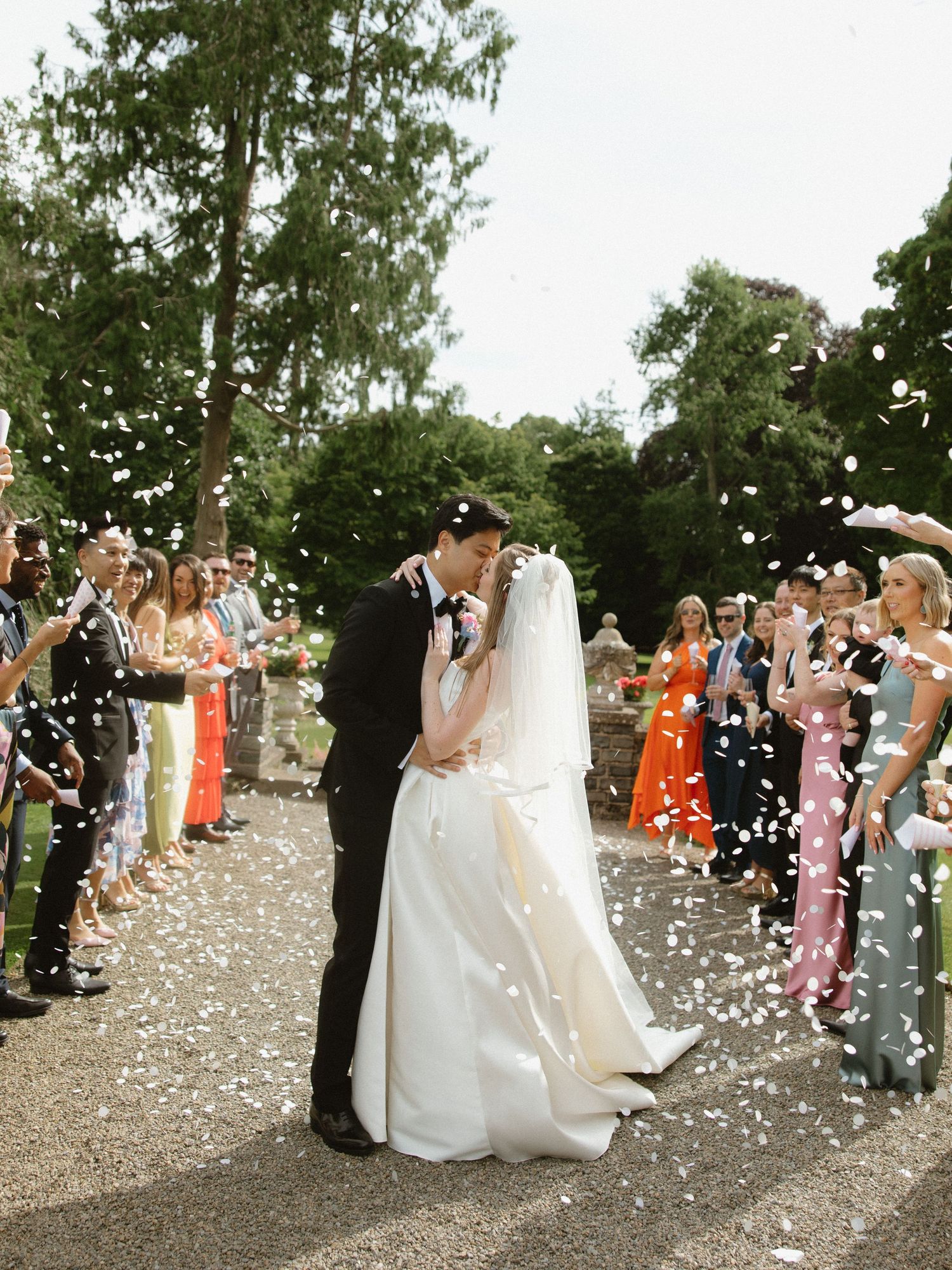 Wedding couple sharing kiss amid shower of confetti from guests on tree-lined path.