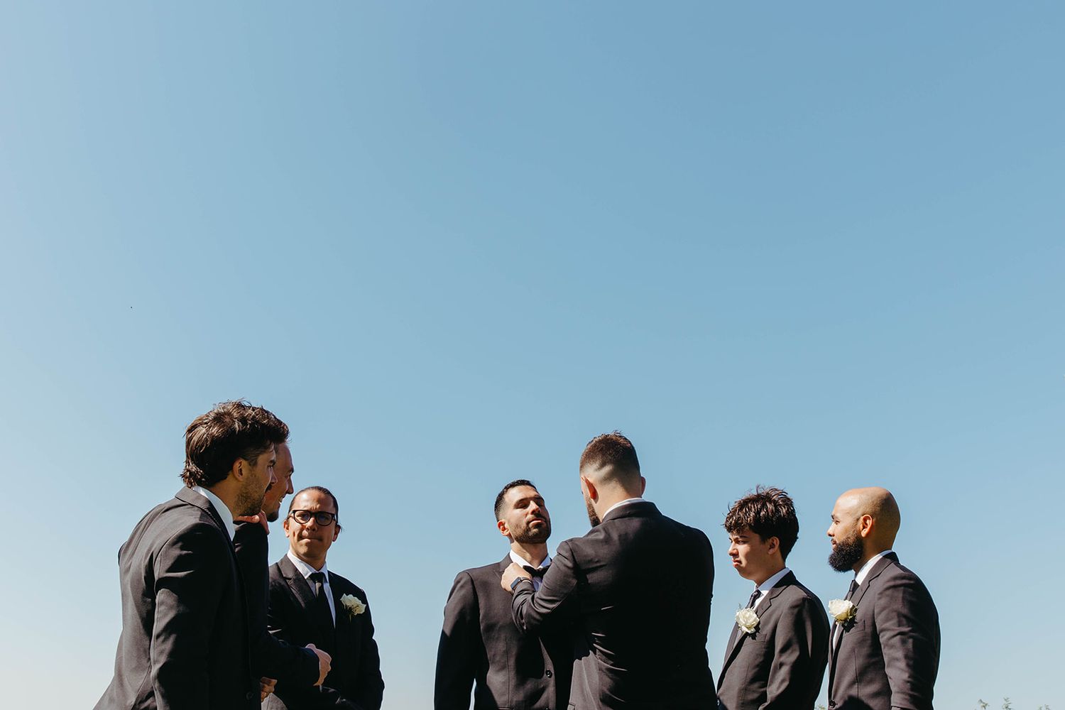 A group of groomsmen in gray suits stand together against a bright blue sky in an outdoor setting.