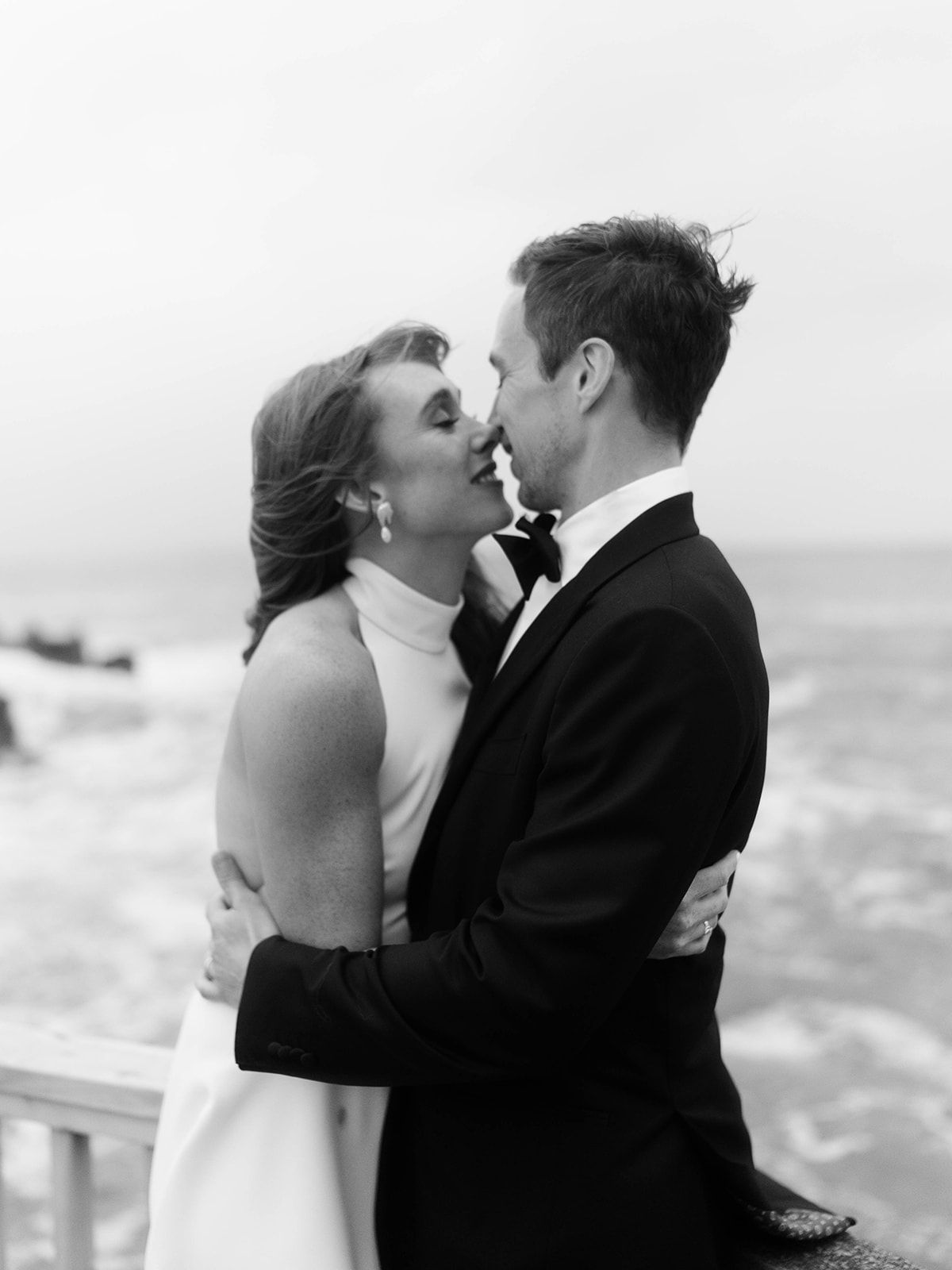 An intimate black and white photo of a couple sharing a kiss on a coastal boardwalk.