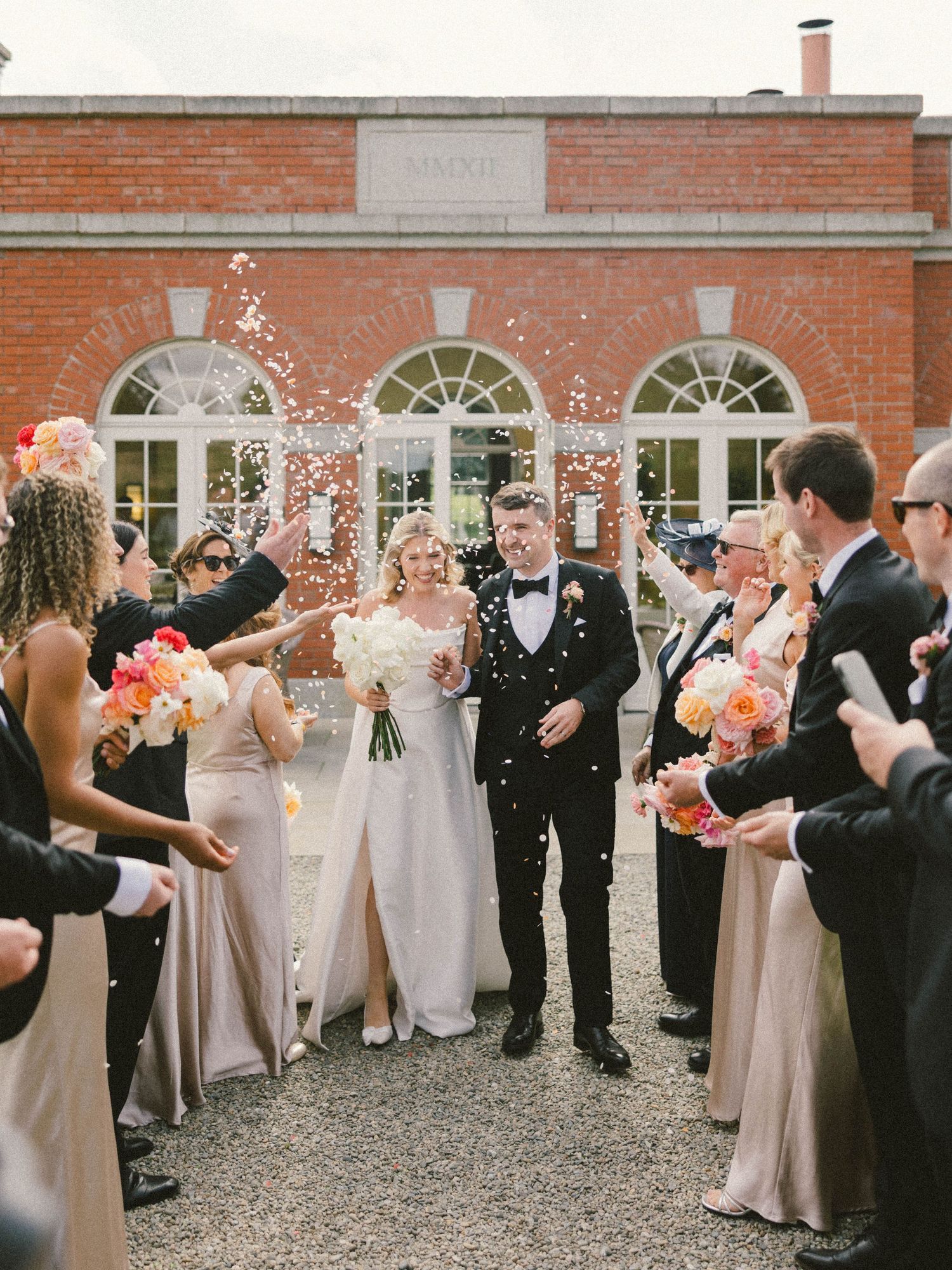 Wedding party throws confetti as newlyweds exit brick building.