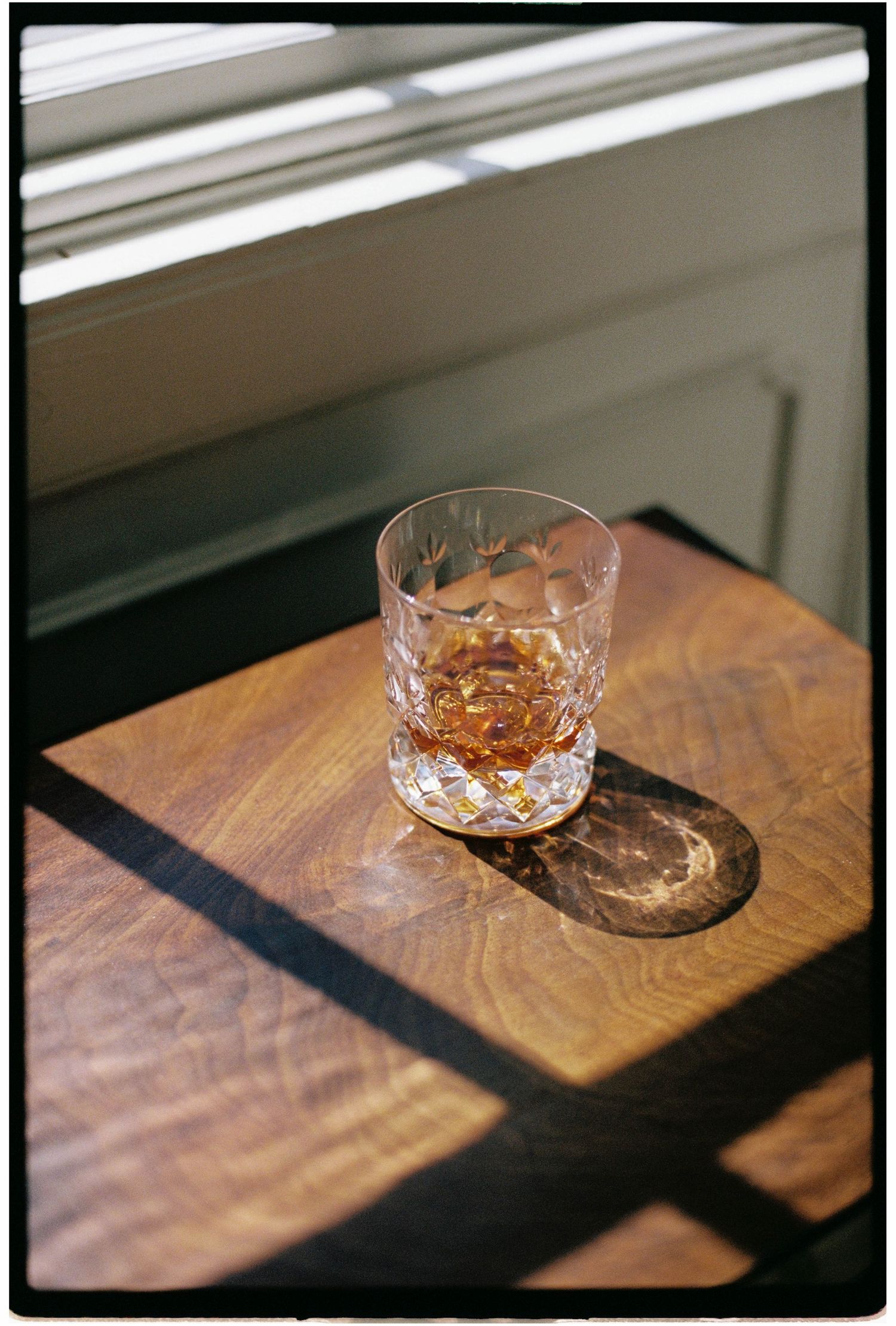 A crystal whiskey glass with amber liquid sits on a wooden table in warm window light casting dramatic shadows.