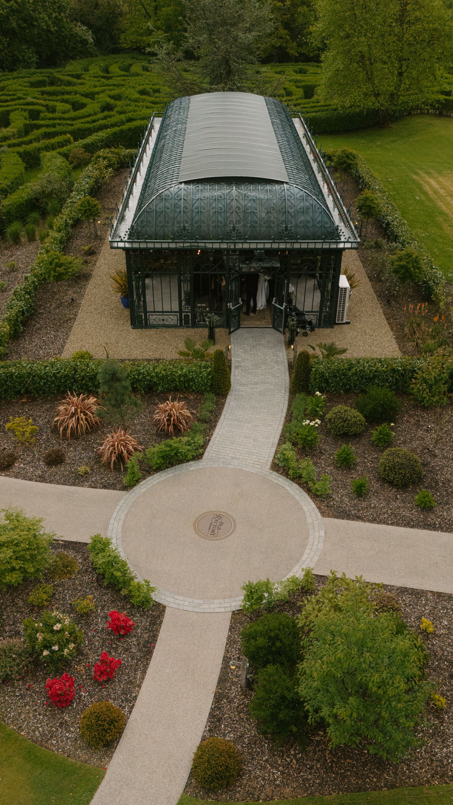 Aerial view of a Victorian greenhouse with curved roof, surrounded by formal gardens, hedgerows, and a circular gravel pathway.