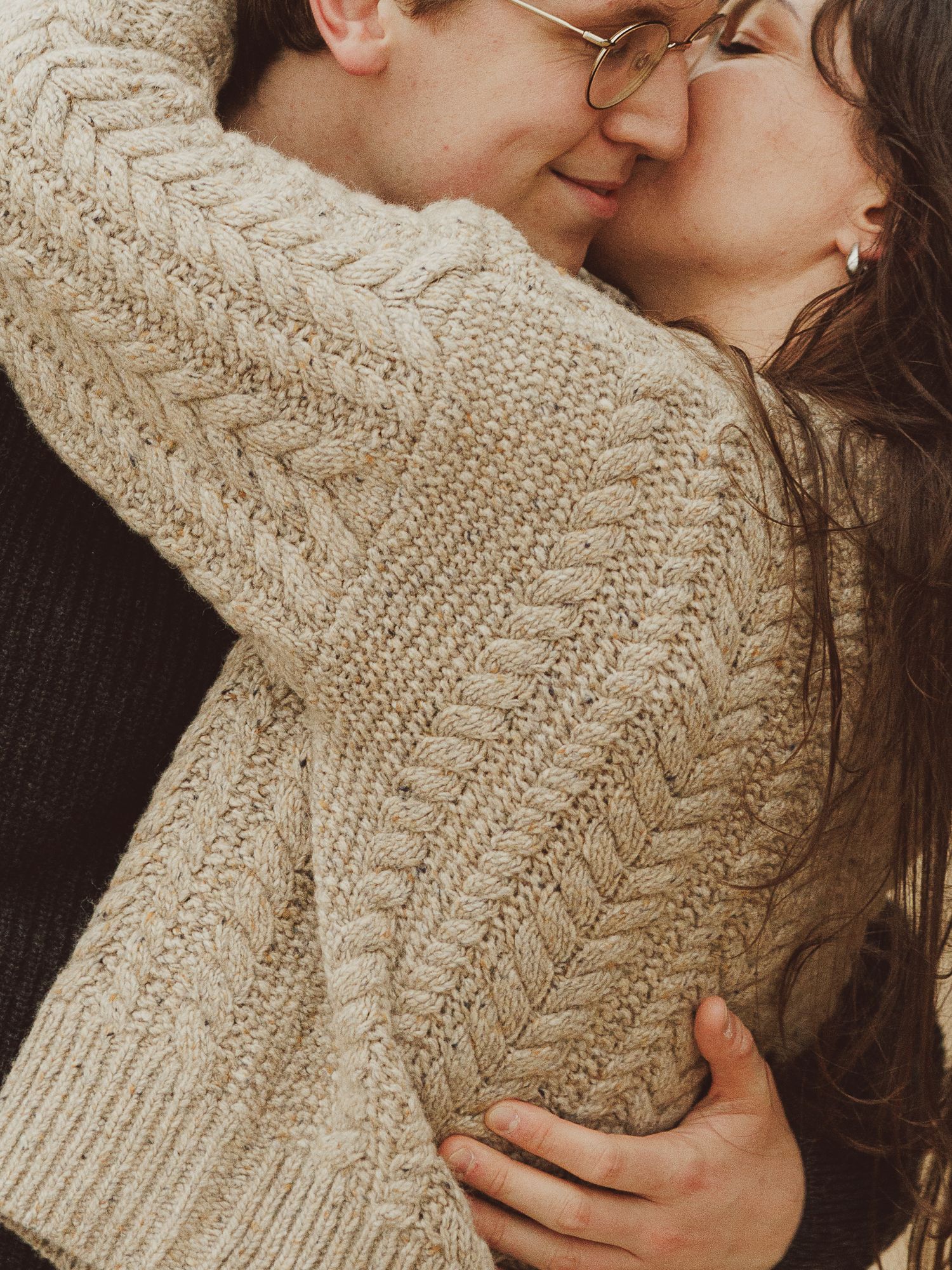 A couple in matching cream cable-knit sweaters share a tender embrace and kiss outdoors.