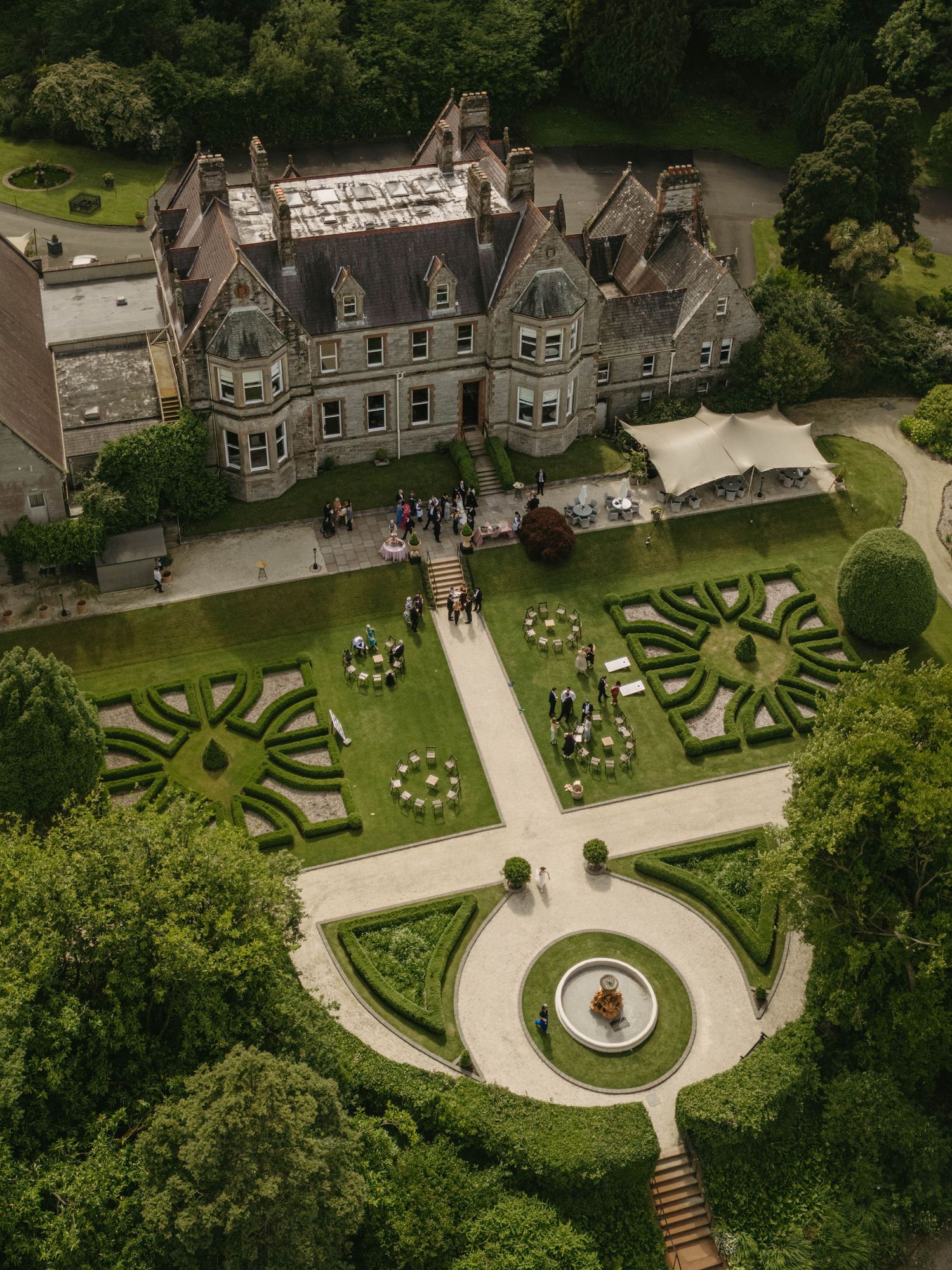Aerial view of a grand Victorian manor with ornate formal gardens, fountain, and wedding guests gathered on manicured grounds.
