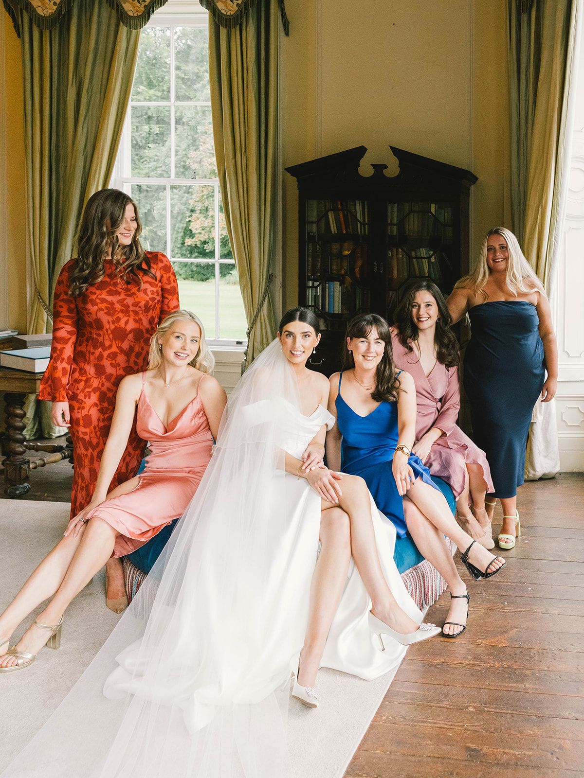 Bridesmaids wearing colorful dresses pose elegantly together in a luxurious room with tall windows and green drapes.