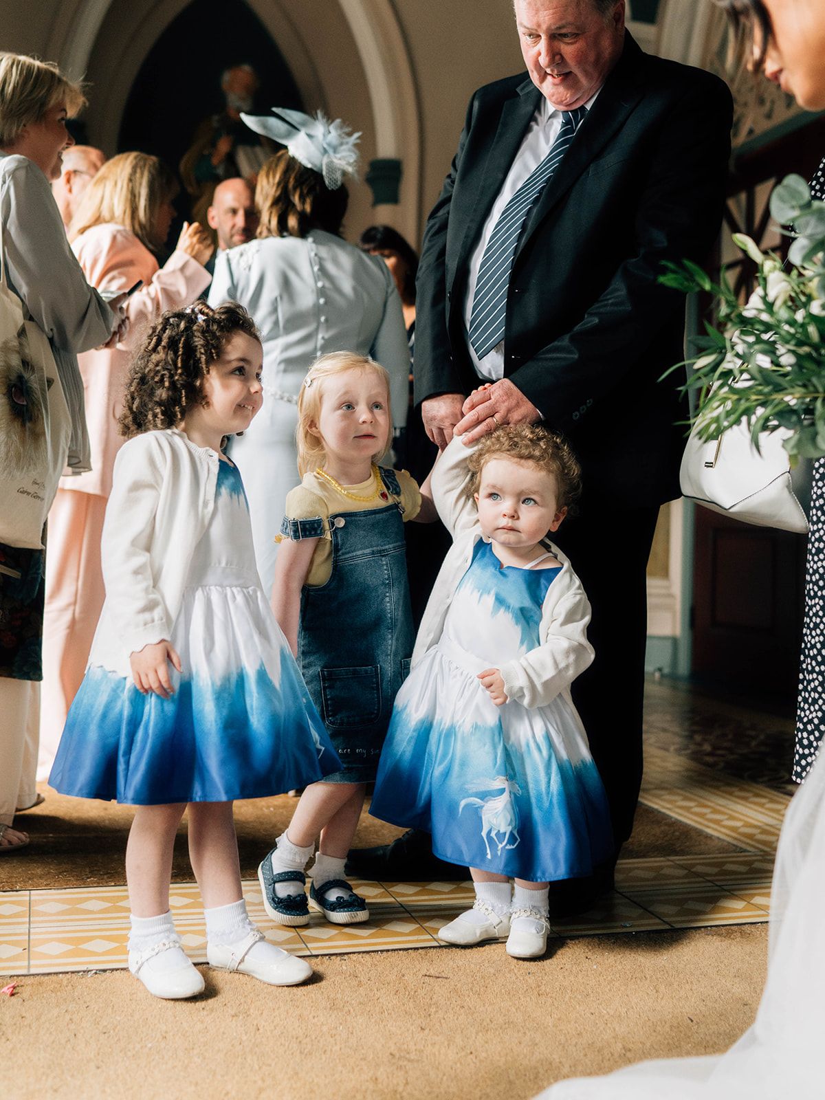 Young children in matching blue and white dresses stand together at a formal event with guests in the background.